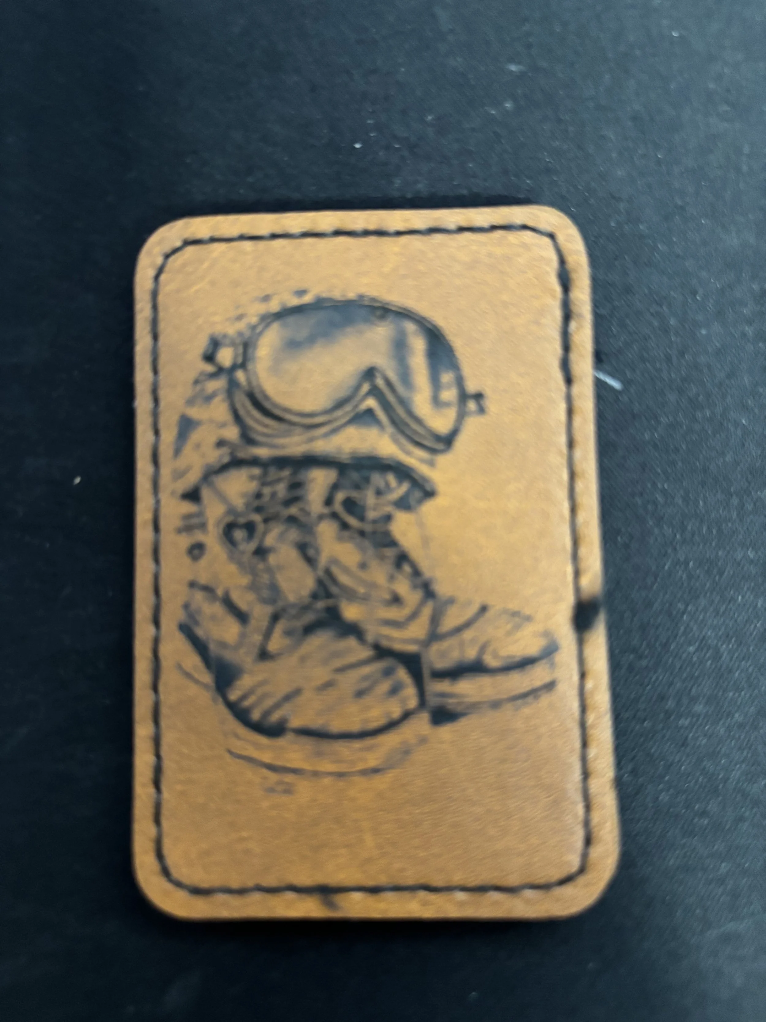 A leather patch with an illustration of a person wearing goggles, a jacket, and slippers, sitting cross-legged.