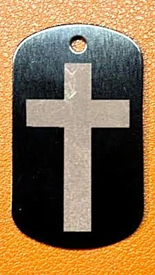 Dog tag with a white cross symbol on a black background, lying on a textured orange surface.