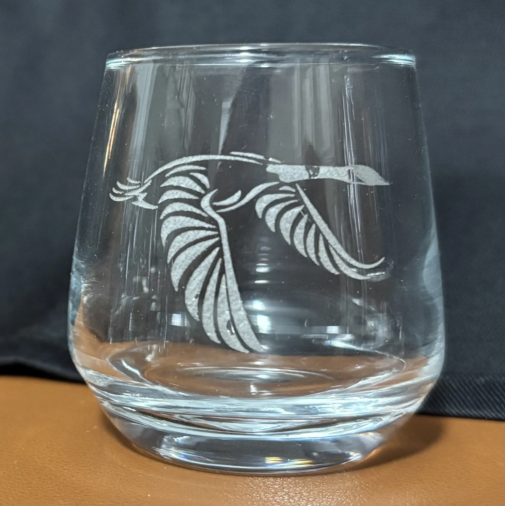 A clear glass with etched design of a heron and tropical leaves, placed on a brown surface with a black background.