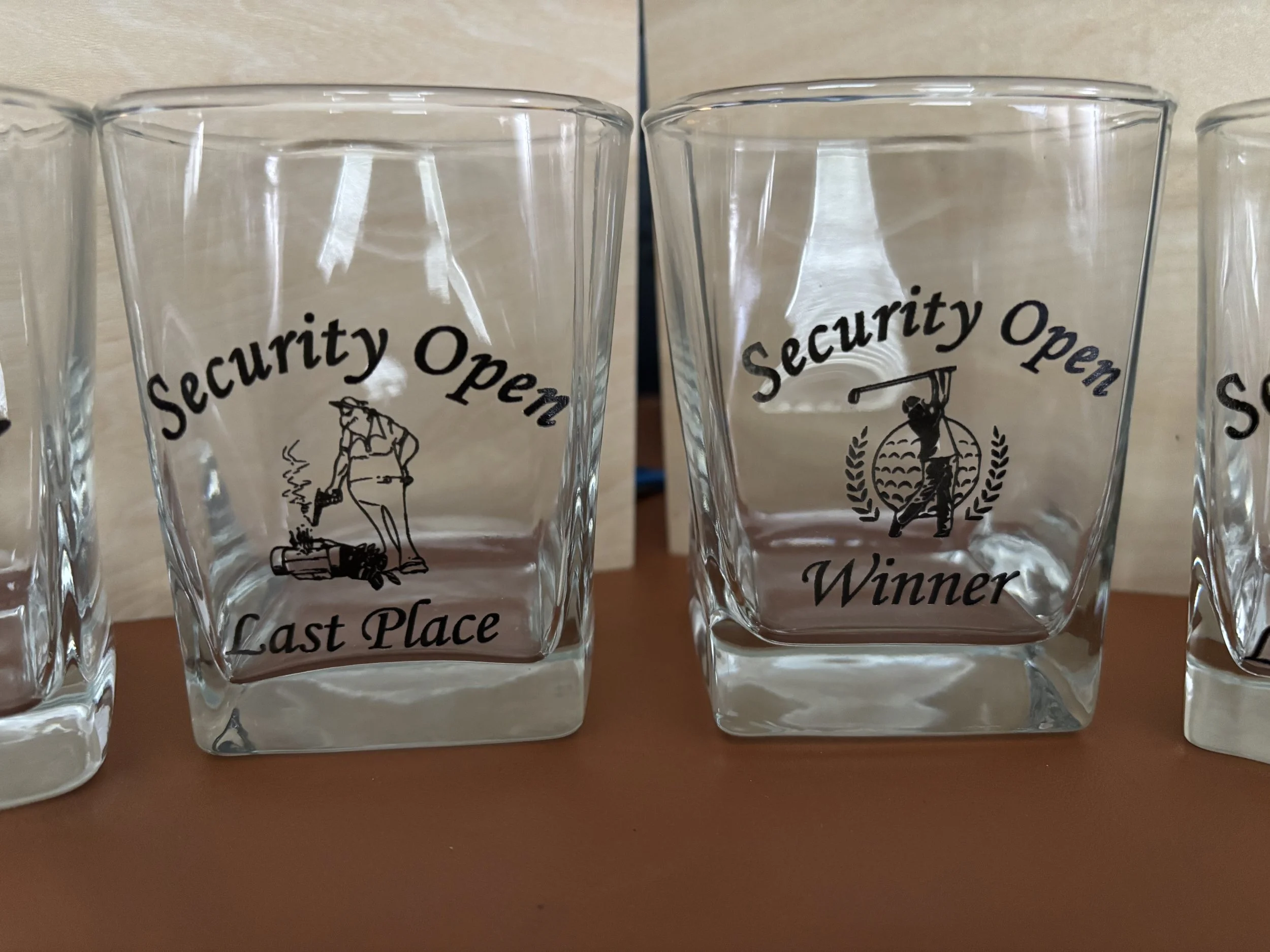 Two glass trophies with engraved golf figures and text, one labeled 'Last Place' and the other 'Winner', both inscribed with 'Security Open'.