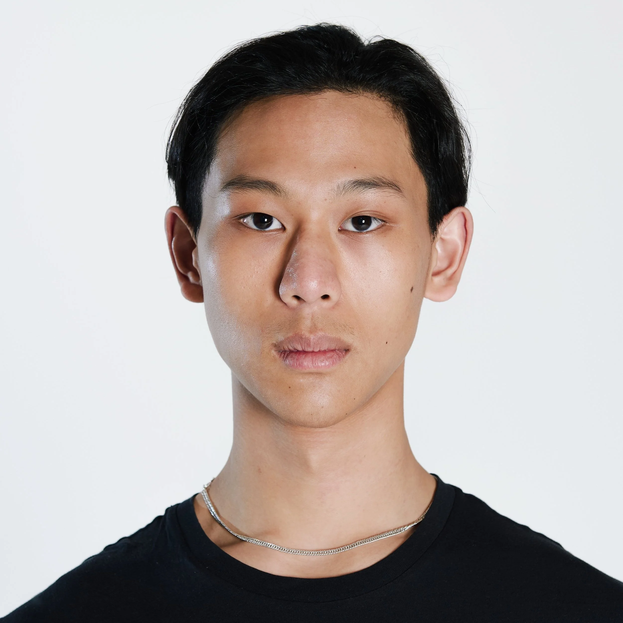 Ragazzo con capelli neri corti, vestito con maglietta nera e collana argento, sfondo bianco.