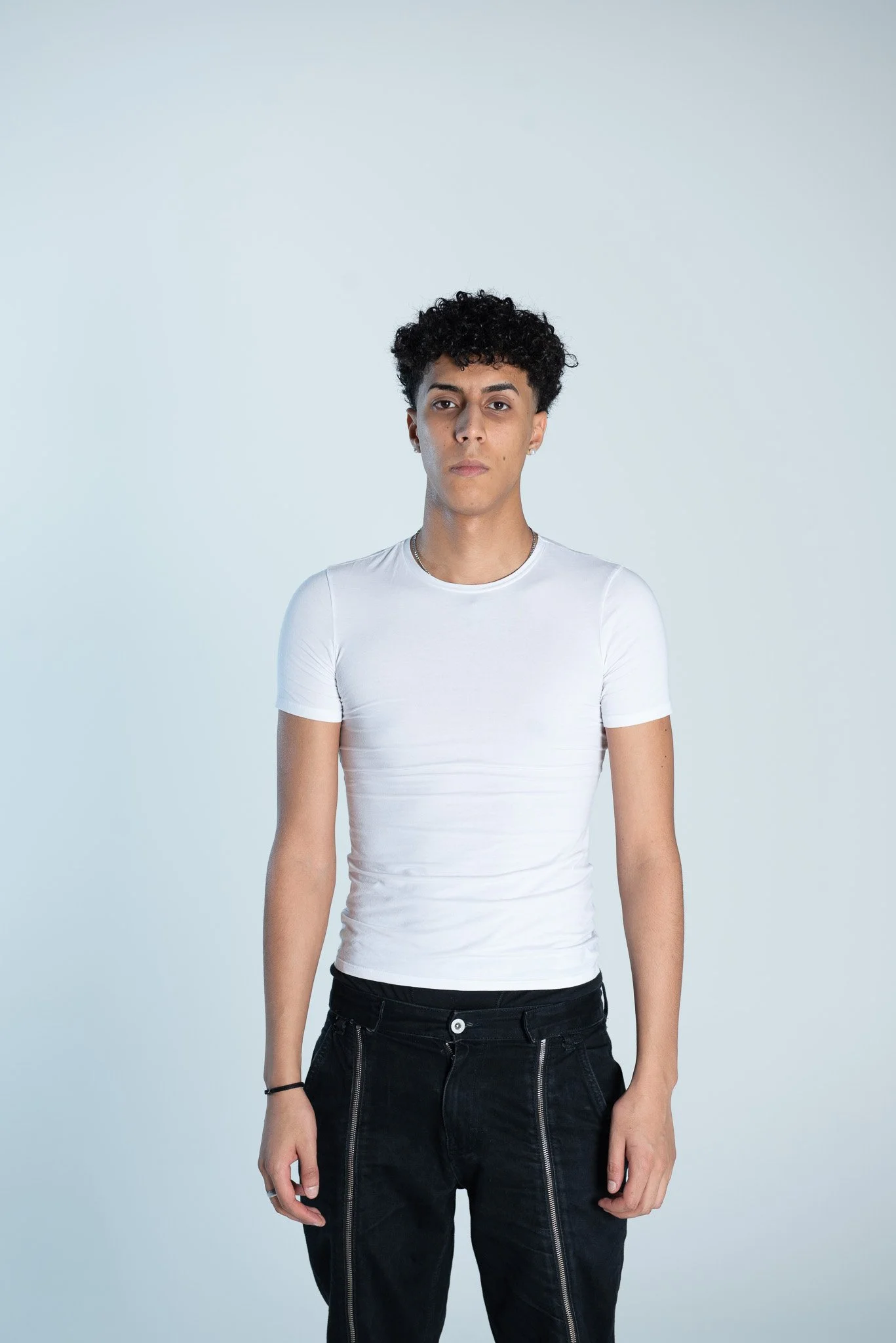 Ragazzo giovane con capelli ricci, vestito con maglietta bianca e pantaloni neri, in piedi con uno sfondo chiaro.