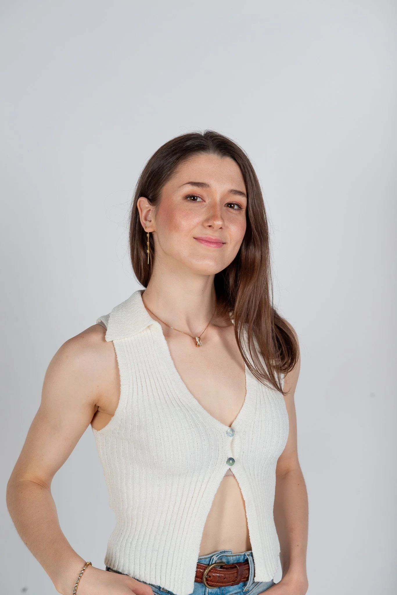 Ragazza con cappottino di maglia bianco, jeans e braccialetti, sorridente davanti a uno sfondo bianco, con orecchini dorati e collana.