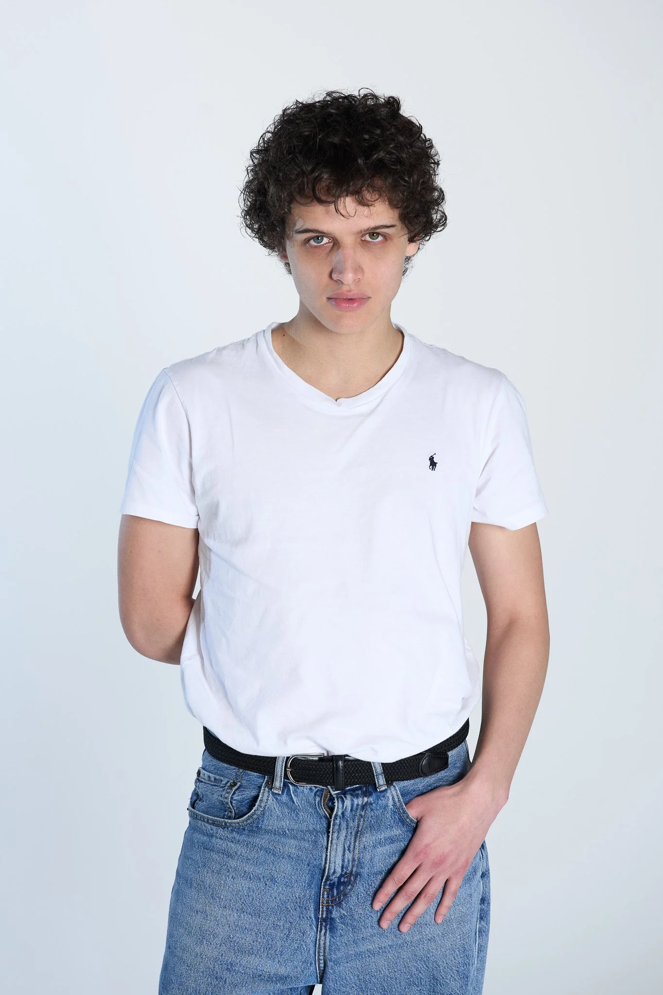 Giovane uomo con capelli curly e occhi chiari, indossa una maglietta bianca con logo di un cavallo e jeans azzurri, sfondo bianco.