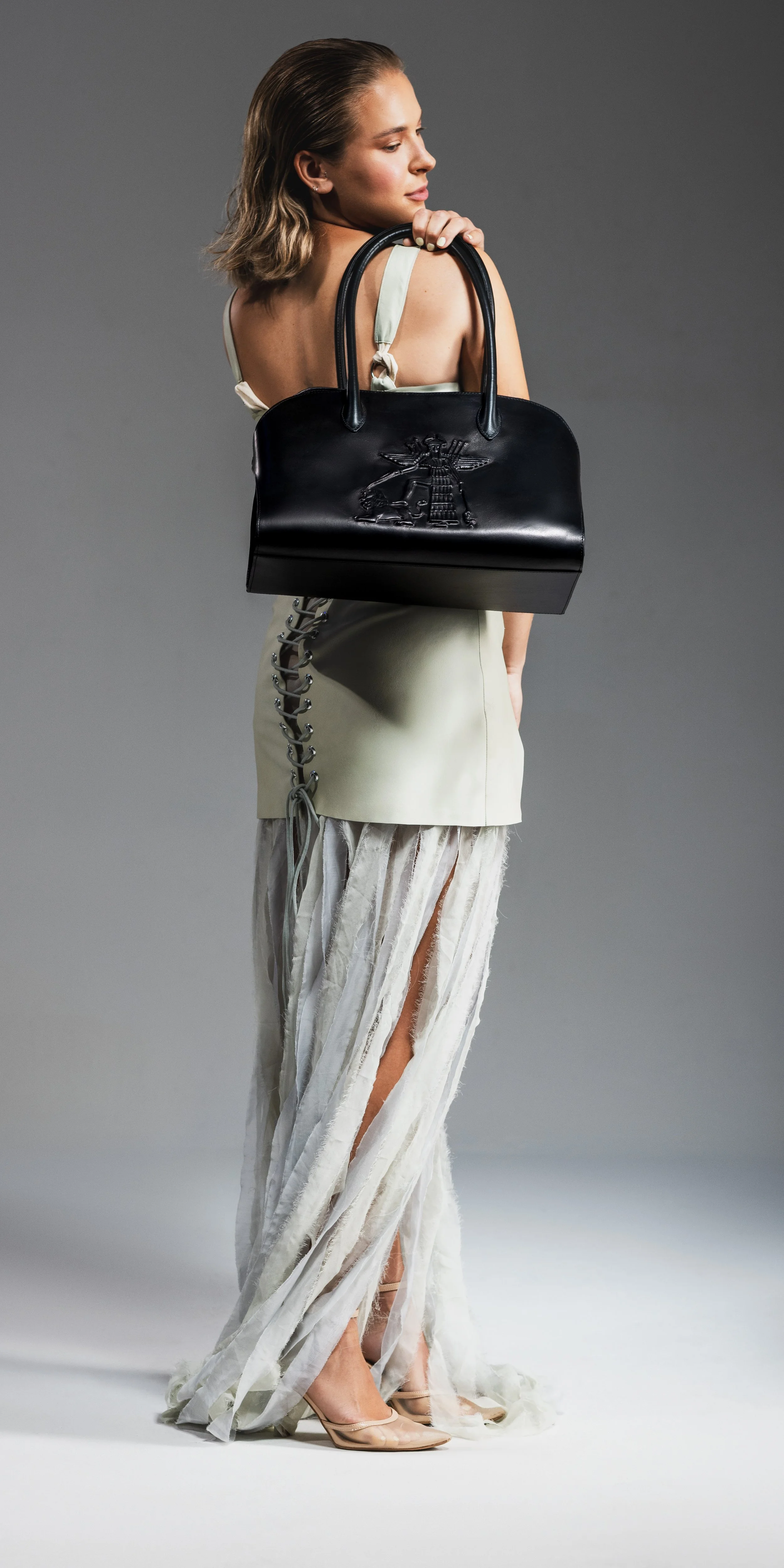 A woman with shoulder-length blonde hair, dressed in a white sleeveless dress with a lace slit, holds a black handbag with embossed designs, standing on a plain gray background.