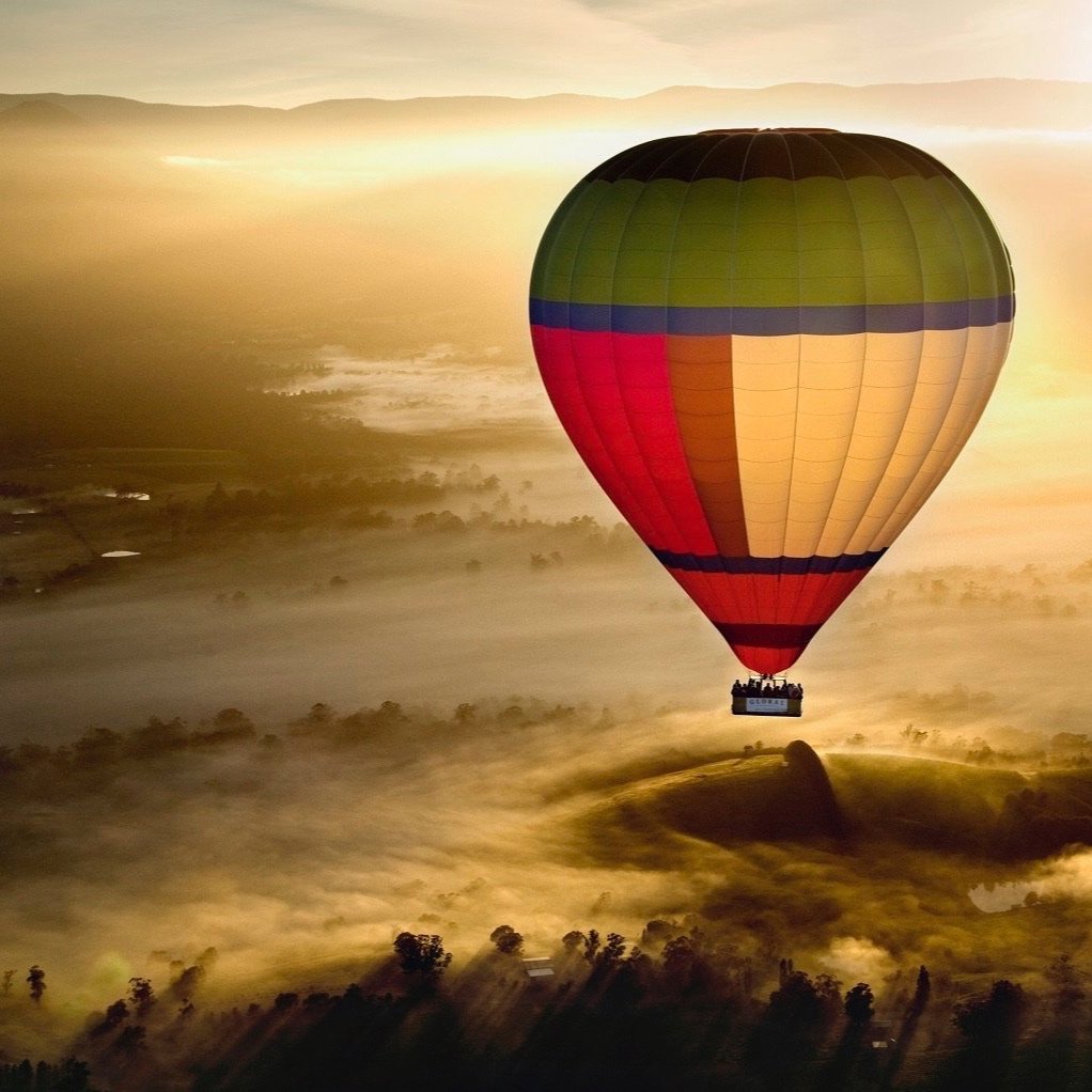 Hot air balloon flying over a foggy landscape at sunrise, with a colorful design and passengers in the basket.