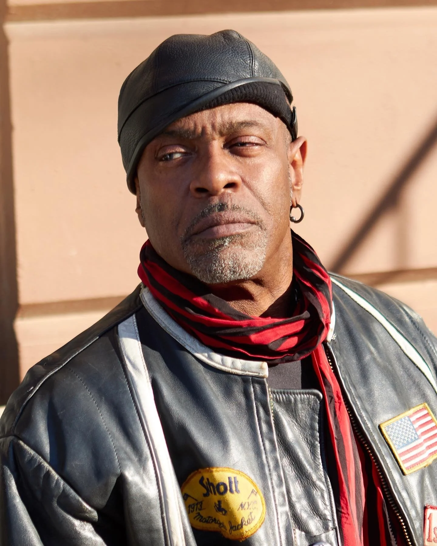 Man wearing a leather cap and jacket with a scarf and earring, standing against a wall.