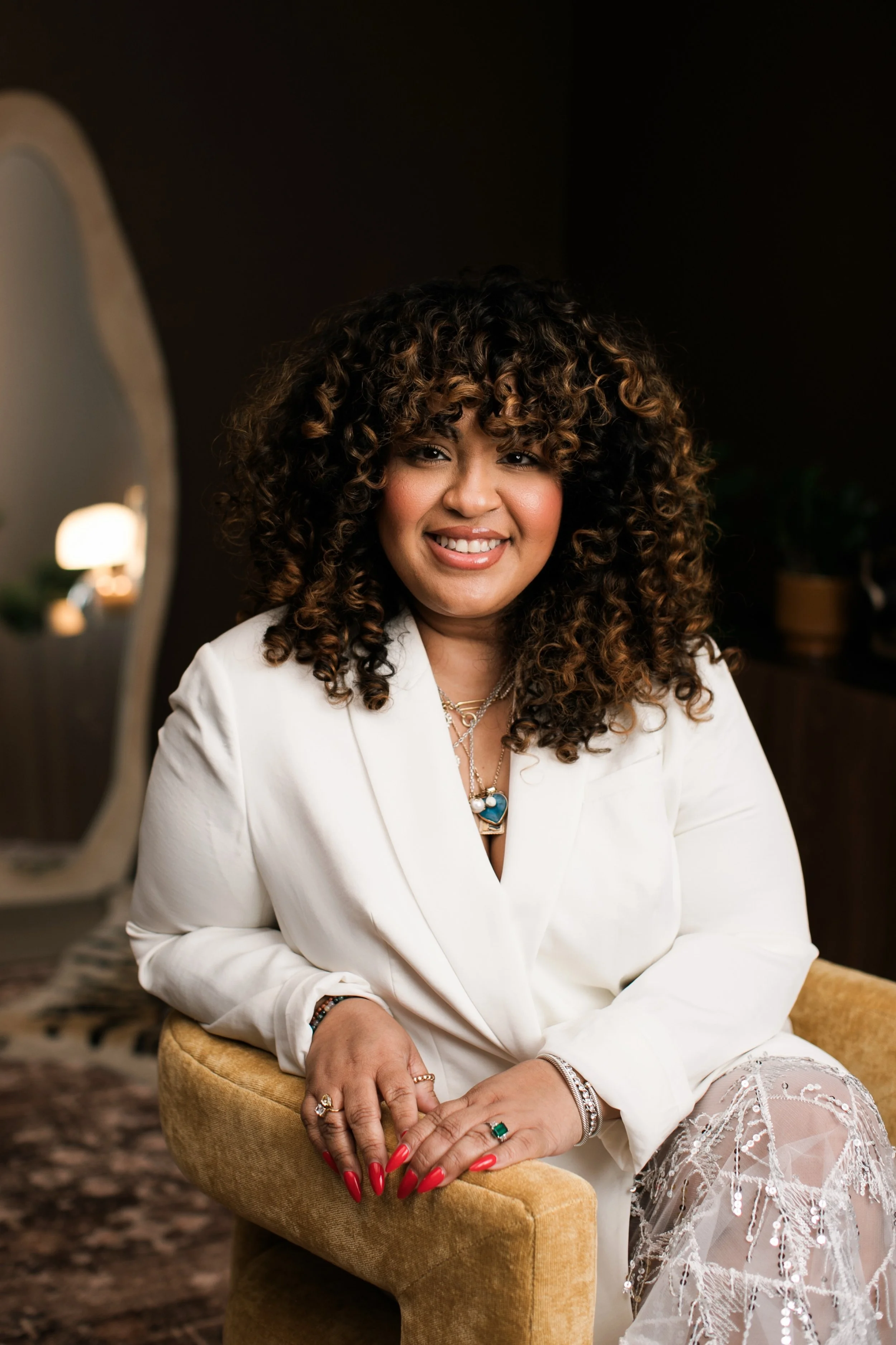 A woman with curly brown hair and wearing a white blazer, rings, and jewelry, sitting on a beige chair and smiling at the camera in a warmly lit room.