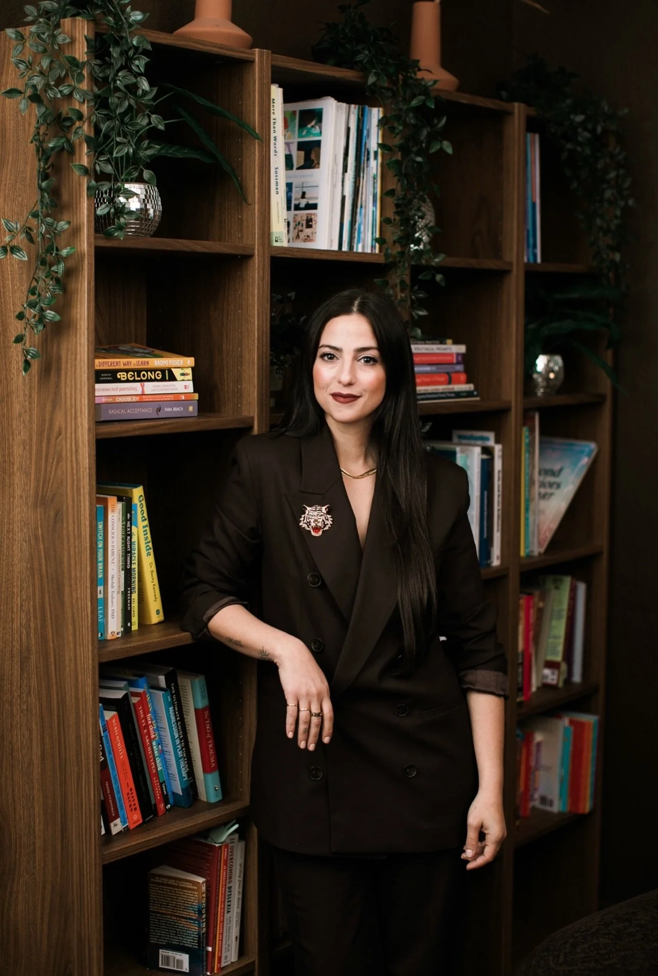 A woman with long black hair, wearing a black blazer with a tiger brooch, is standing in front of a wooden bookshelf filled with books and decorative items, including plants and vases.