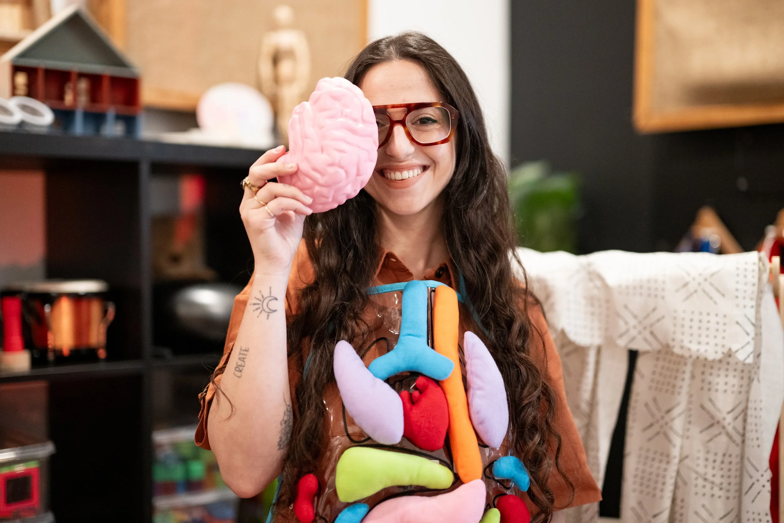 Woman wearing glasses holding a pink brain model over one eye, dressed in an apron with colorful anatomical organ designs, smiling in a room with shelves and crafts.