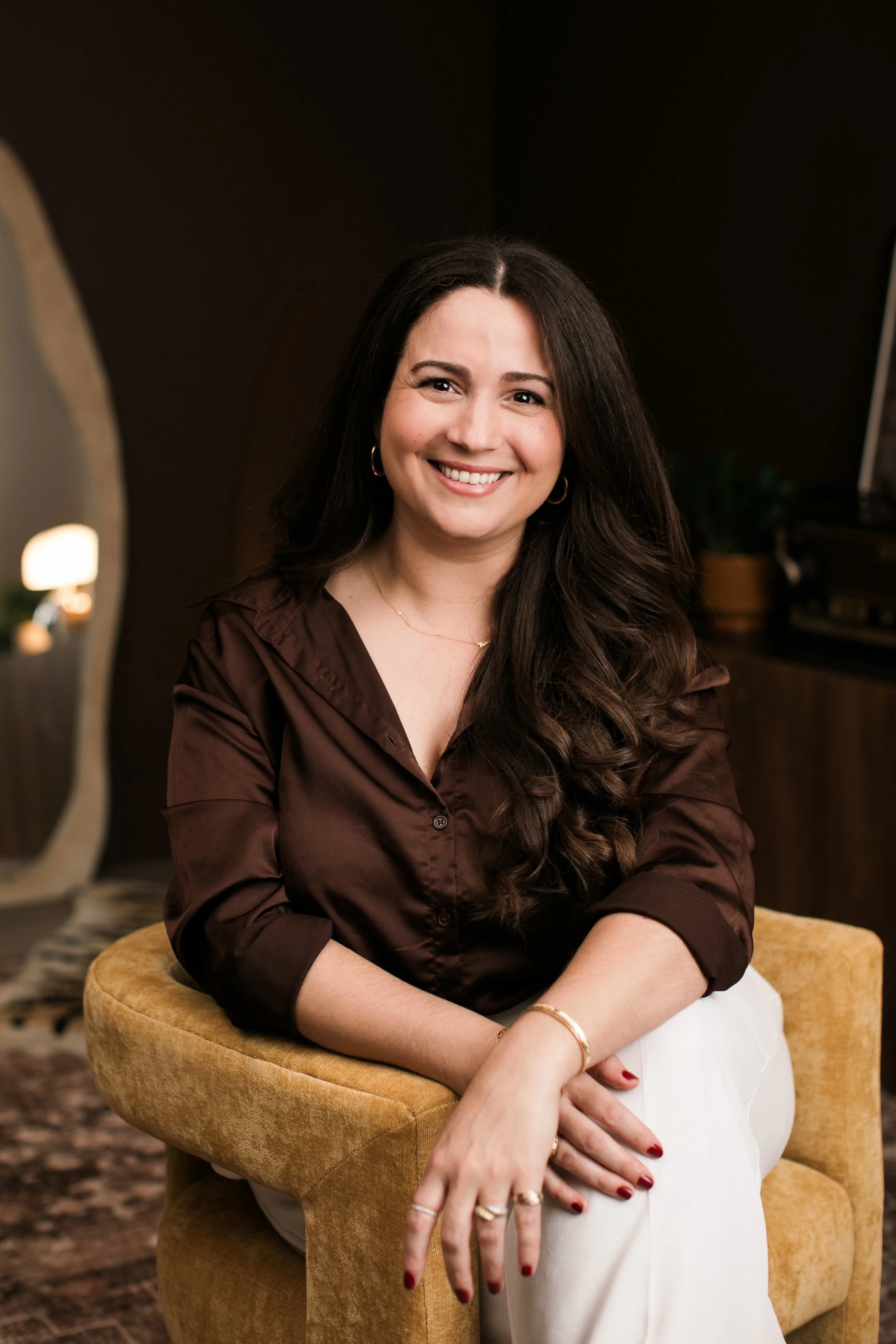 A woman with long dark hair smiling while sitting on a yellow armchair in a cozy, warmly-lit room.