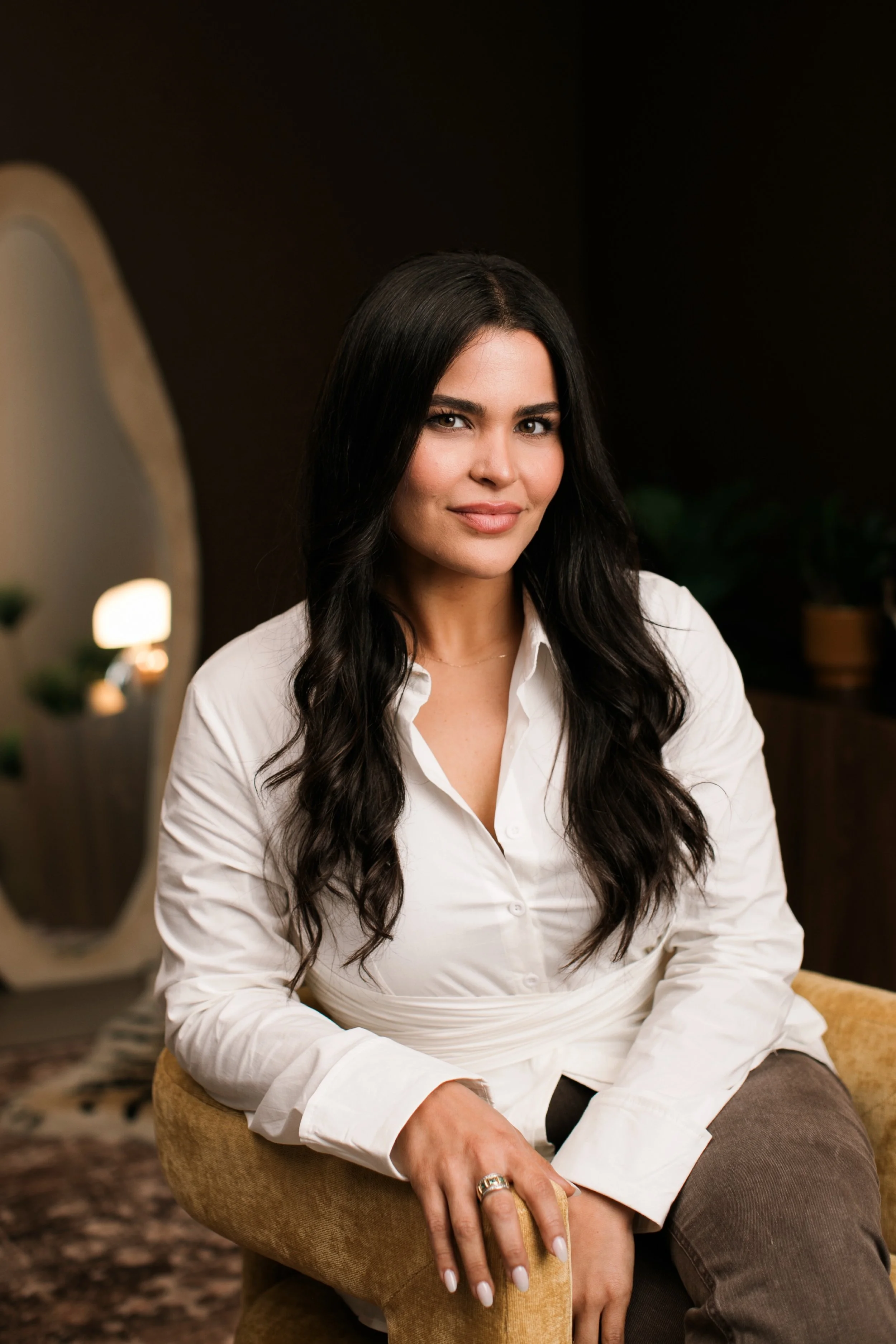 A woman with long black hair and a white shirt sitting on a yellow chair in a dimly lit room.