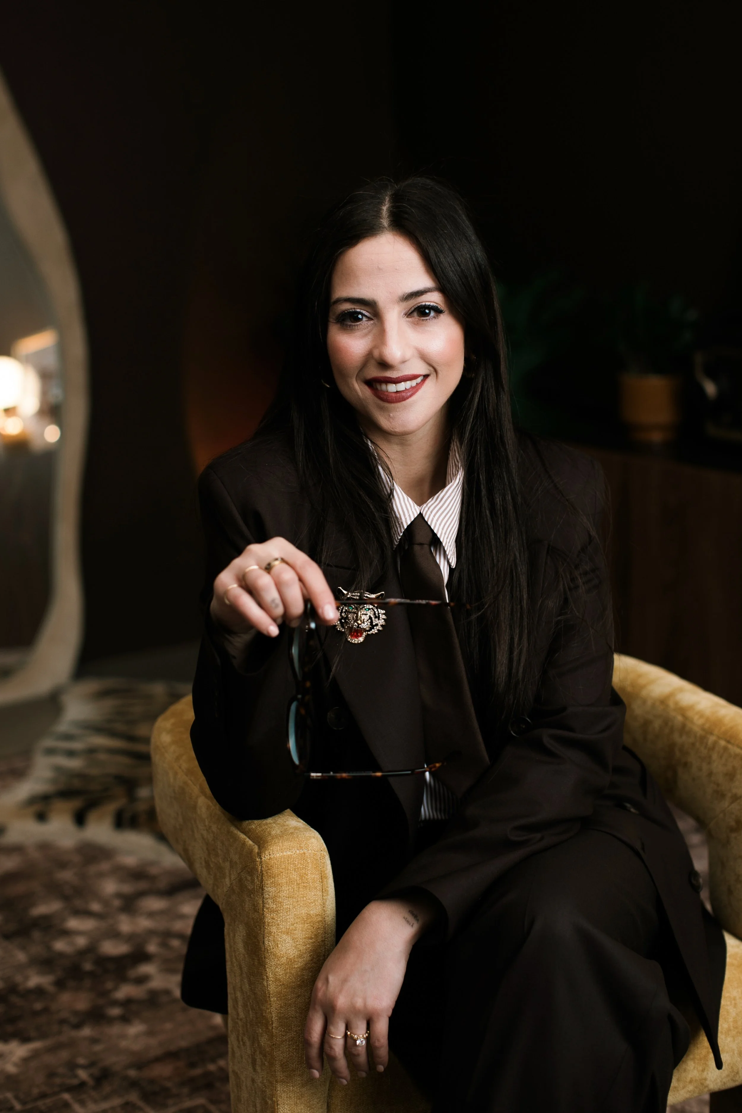 A woman with long dark hair sitting in a yellow armchair, wearing a dark blazer and holding a pair of glasses, smiling at the camera.