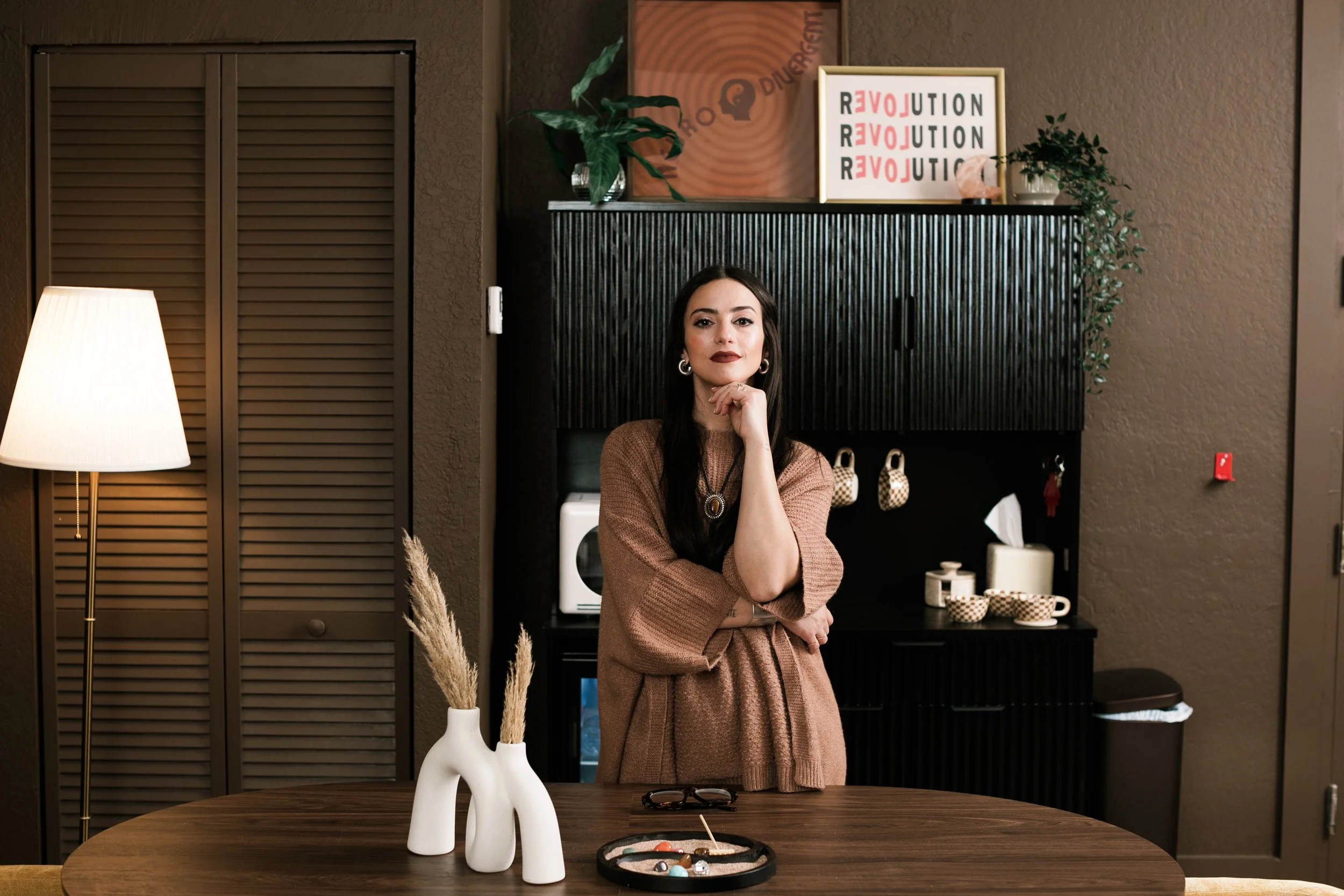 A woman with long dark hair, wearing a brown dress, standing with her hand on her chin in a modern, warmly lit room with decorative items and framed art on the wall.