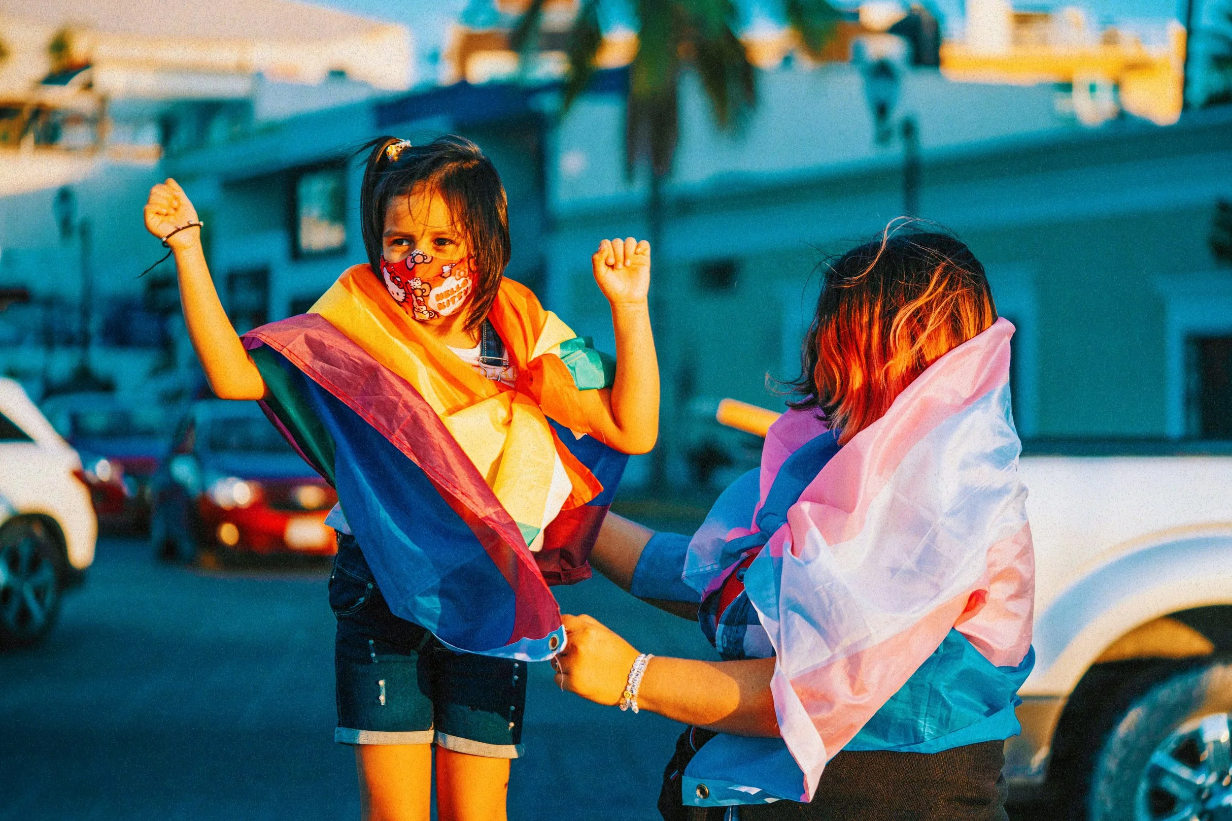 Niña con bandera del arcoíris y mascarilla celebra con otra persona en la calle, con coches y edificios al fondo.