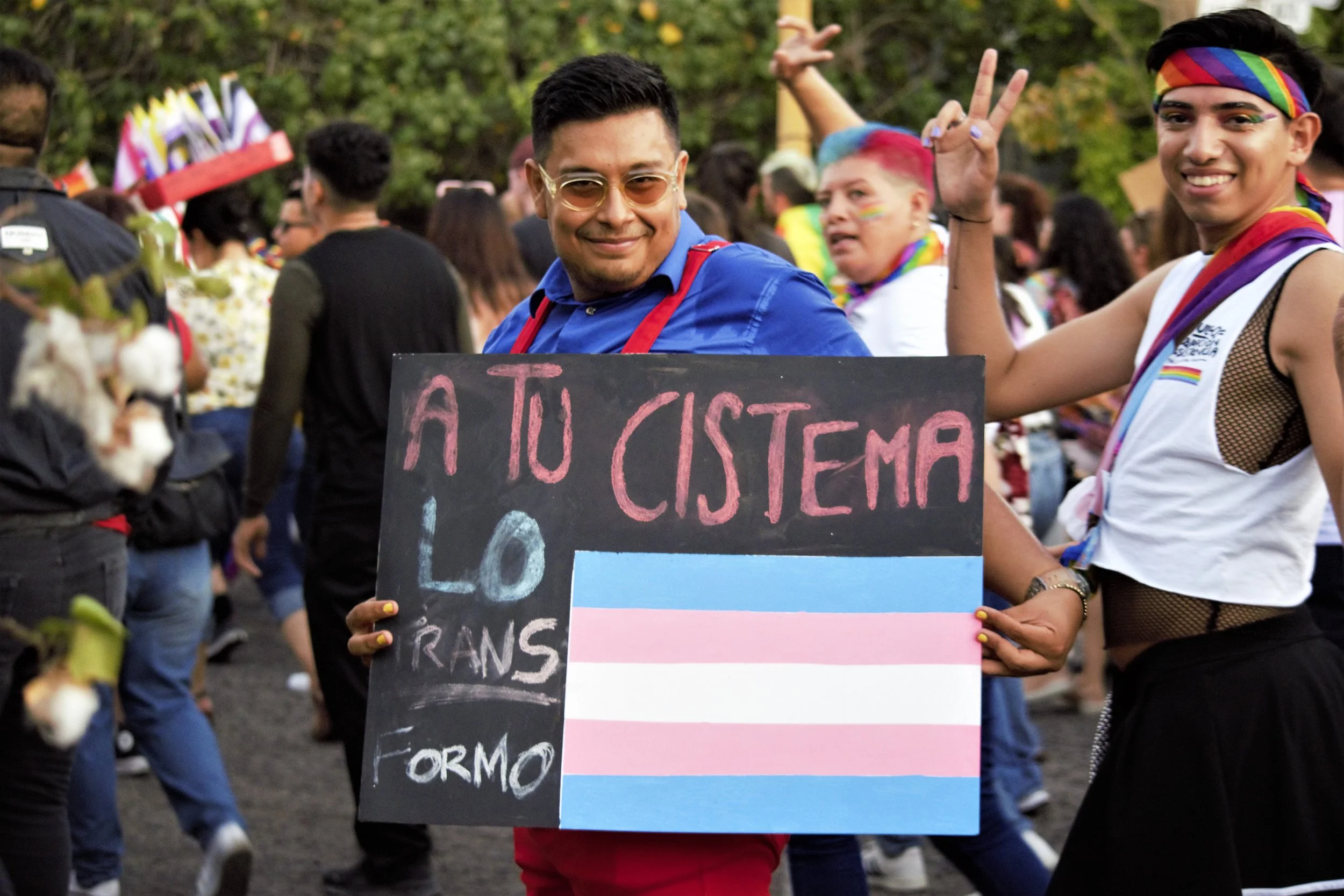 Persona en un evento del orgullo LGBTQ+ sosteniendo cartel que dice "A tu cisterna, lo transFormo" junto a la bandera transgénero.