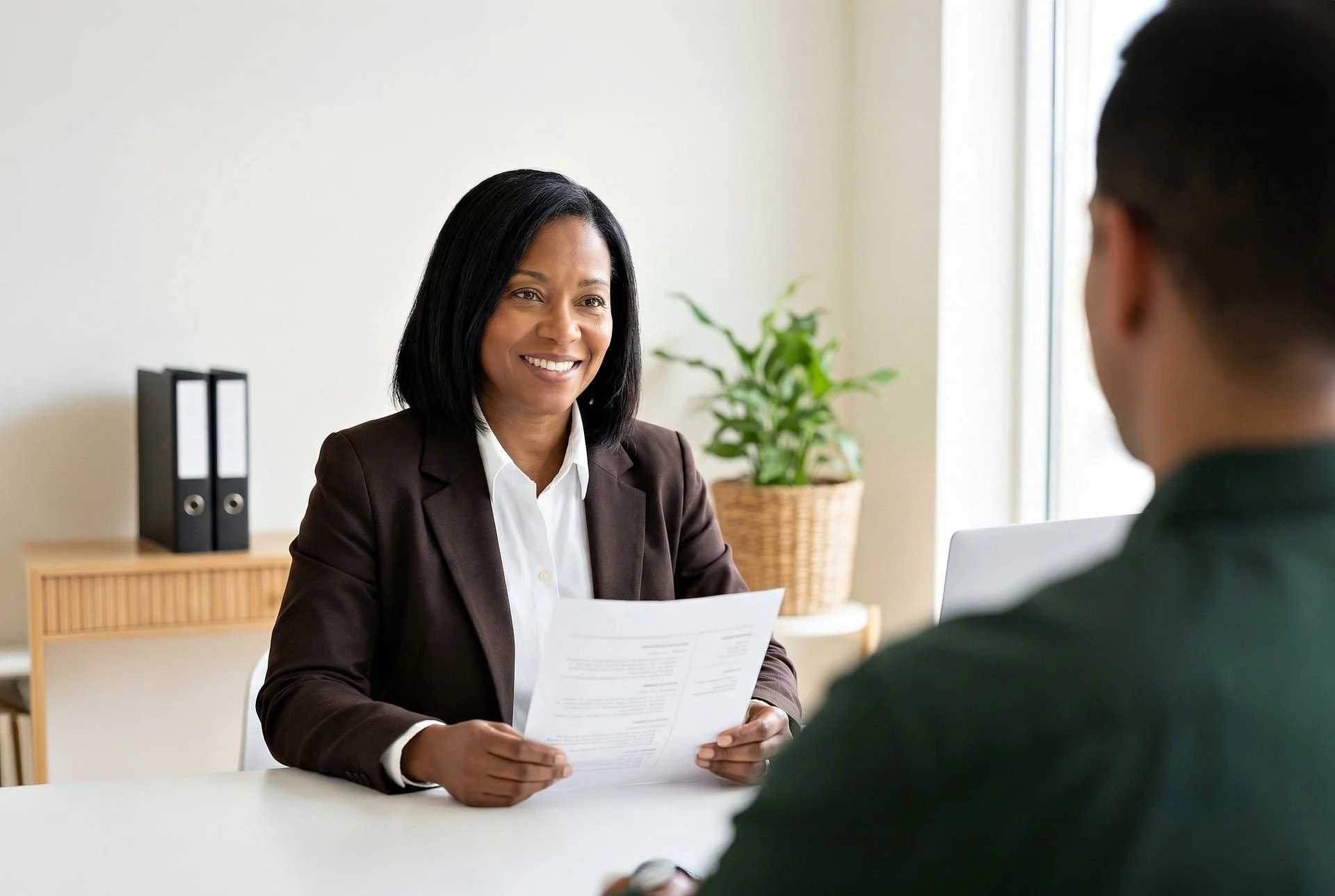 A woman in a brown blazer smiling and holding a document, sitting across from a person in a black blazer, in a bright office with a potted plant and black binders in the background.