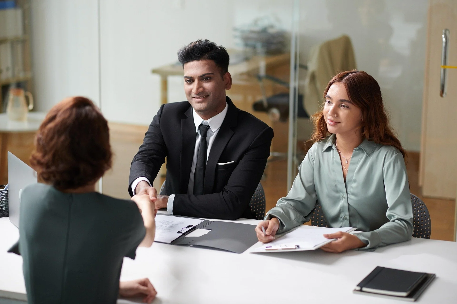 A professional business meeting with three women and one man. The man, dressed in a black suit with a tie, is shaking hands with an older woman with short hair, while a young woman with long red hair, dressed in a light gray shirt, looks on. The setting appears to be an office environment with documents, notebooks, and electronic devices on the table.