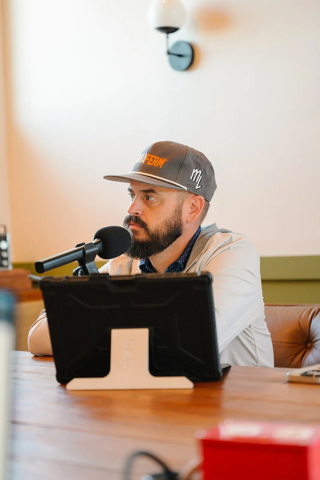 A man with a beard wearing a gray baseball cap with orange and white text, sitting at a table with a microphone and a tablet, in a room with a white wall and modern wall light.