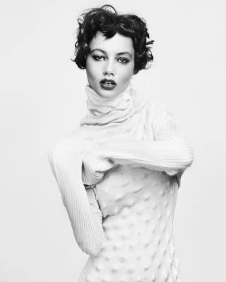 Black and white photo of person with short curly hair wearing a textured turtleneck sweater, posing with hands crossed at the waist.