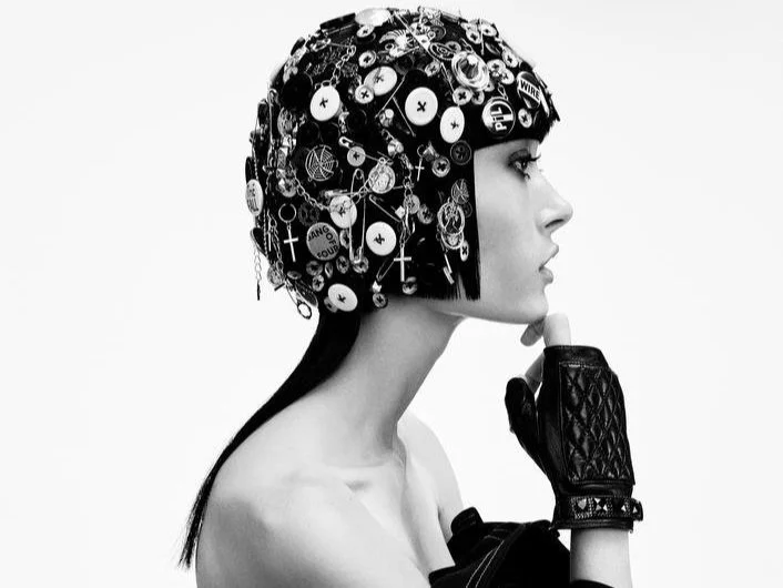 Profile of a person wearing a decorated helmet with buttons and charms, black gloves, black and white photo.