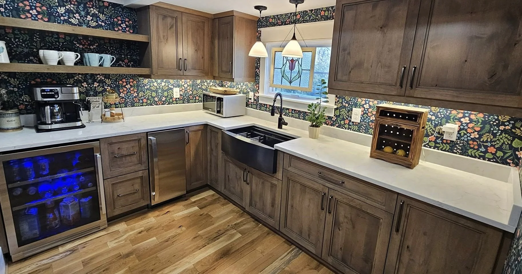 A cozy kitchen with wooden cabinets, a white marble counter, and a colorful floral wallpaper backsplash. It features a microwave, a coffee maker, a small wine cooler, and a decorative wine rack. There's a window above the sink with a stained glass de