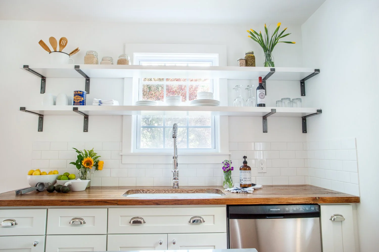 Kitchen with white cabinets, wooden countertop, open white shelves with glass jars, dishes, a bottle of wine, and a sunflower in a vase, window with sunlight, sink with a modern faucet, and a dishwasher.