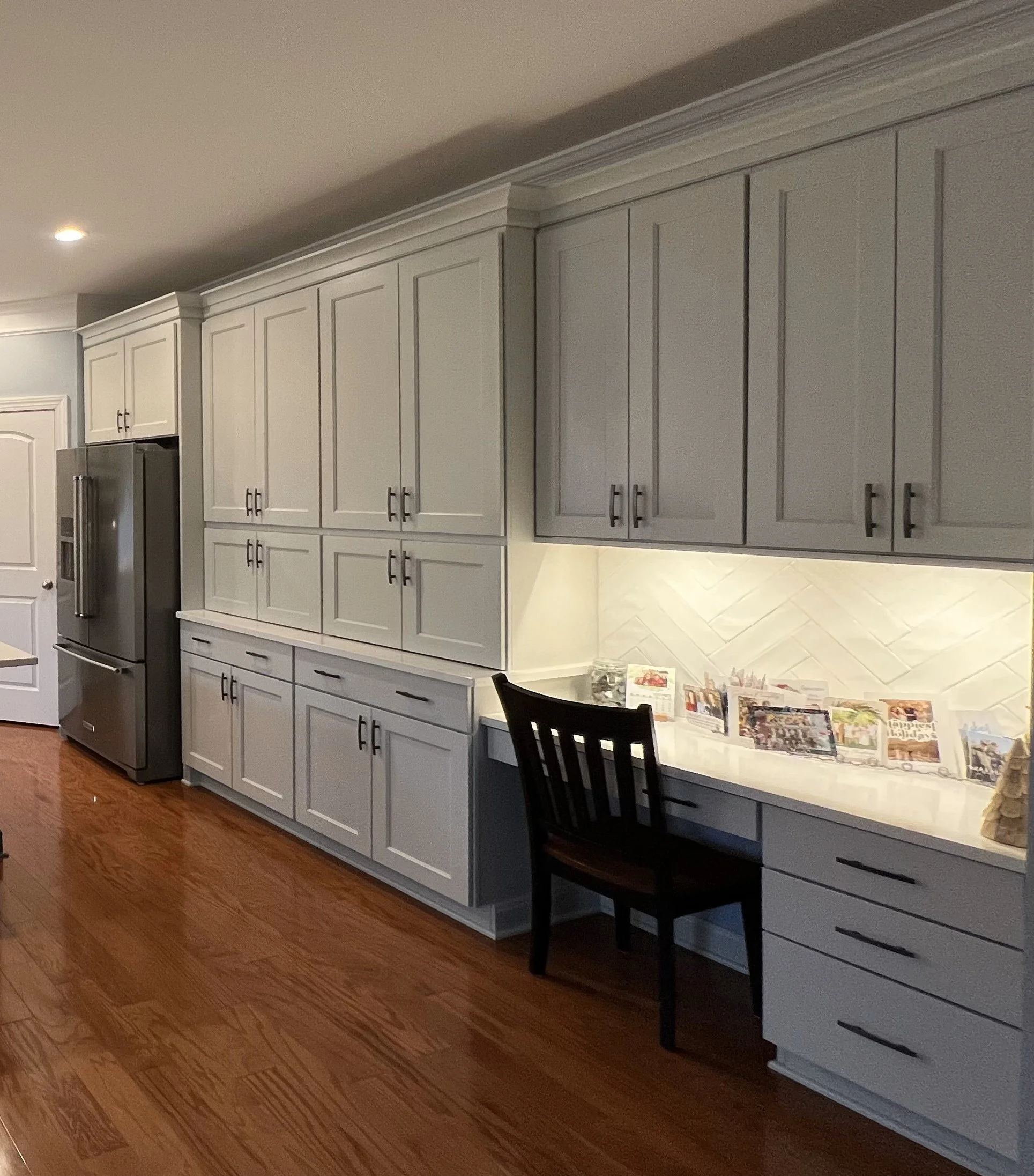 Kitchen with white cabinets, stainless steel refrigerator, hardwood floor, and a small workspace area with a black chair and gathered photographs.