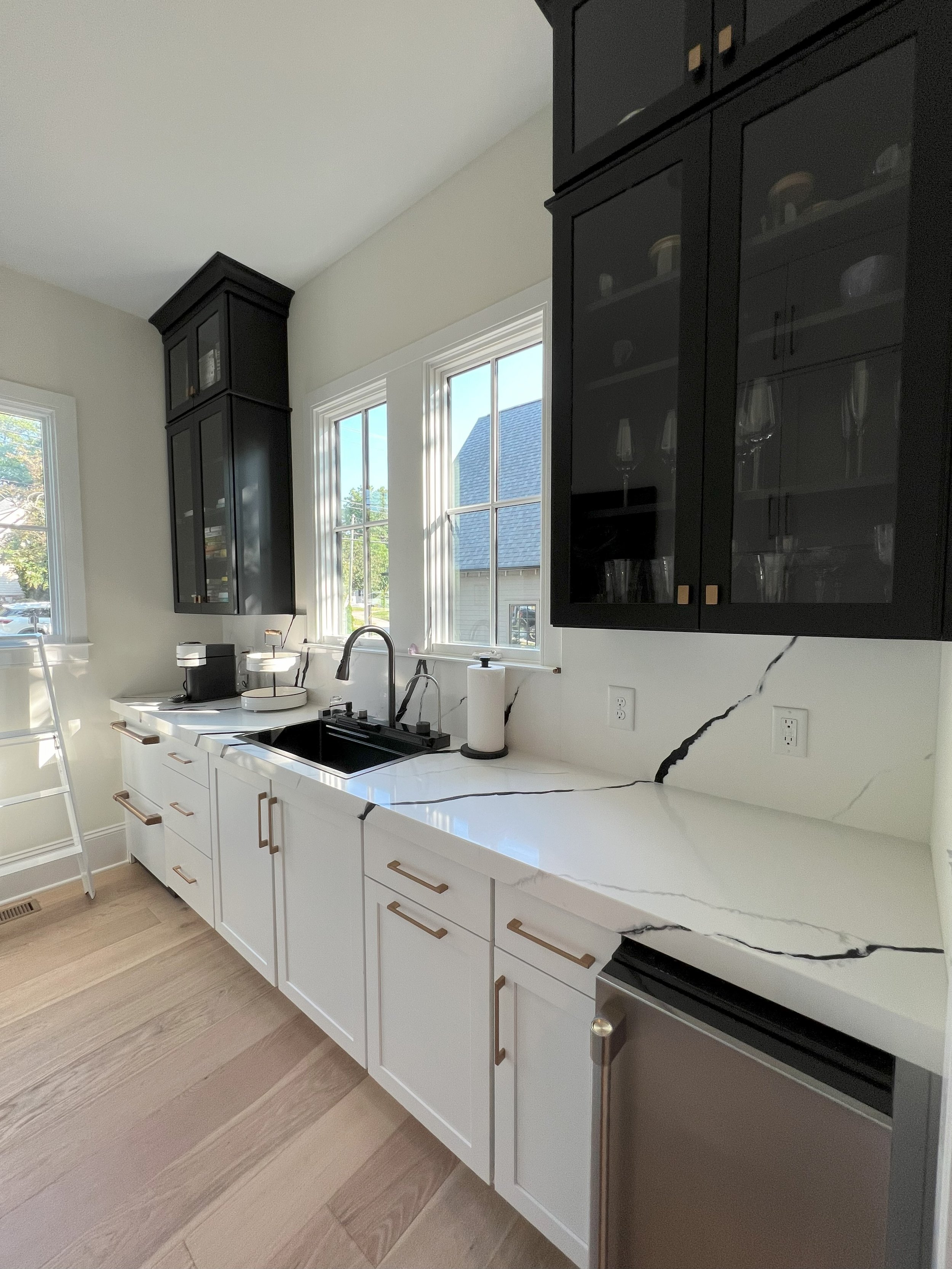 Modern kitchen with white cabinets, black upper cabinets with glass doors, gold handles, a white marble countertop with black veining, and two windows atop the counter letting in natural light.