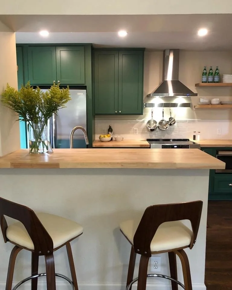A modern kitchen with green cabinets, stainless steel appliances, open shelving with bottles and dishes, a wooden island with a vase of yellow flowers, and two stylish chairs with wood frames and cream cushions.