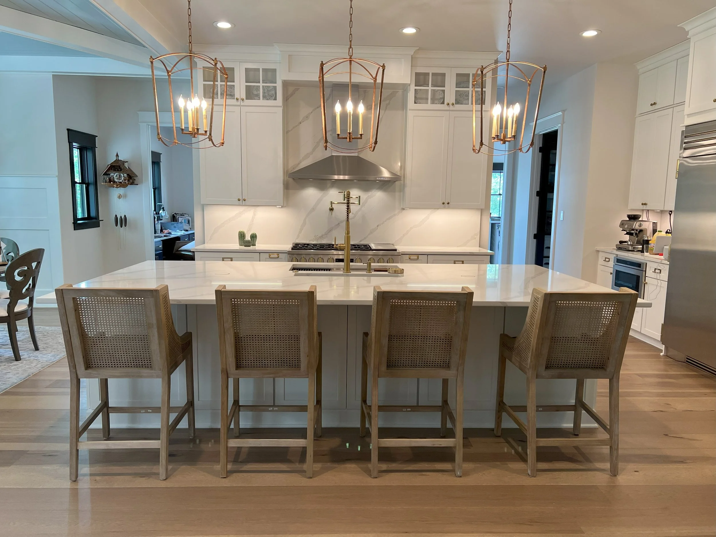 Modern white kitchen with a large island, beige chairs, and gold light fixtures hanging above.