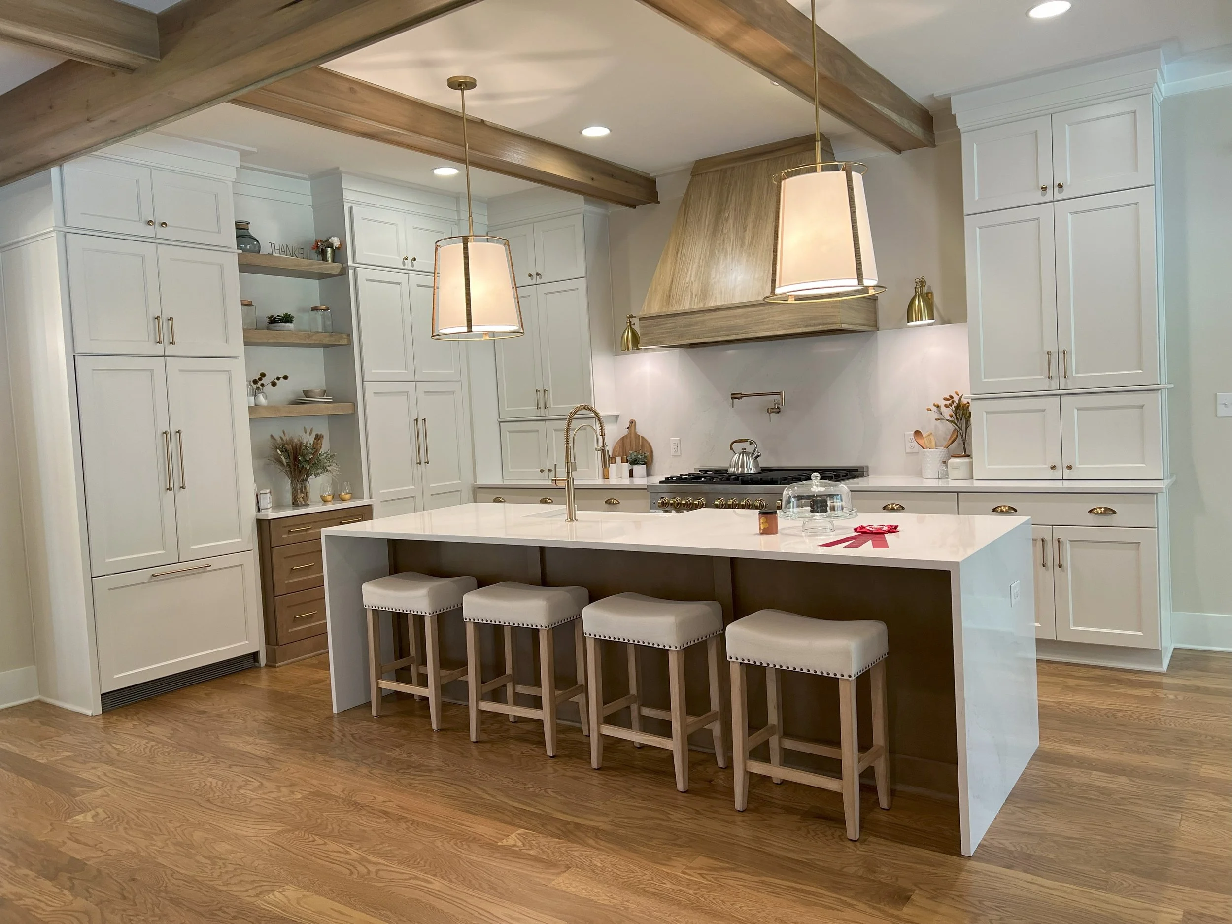 Modern kitchen with white cabinets, wooden accents, a large white island with four beige barstools, and pendant lights hanging over the island. There are open shelves with decorative items and a stove with a wooden vent hood.