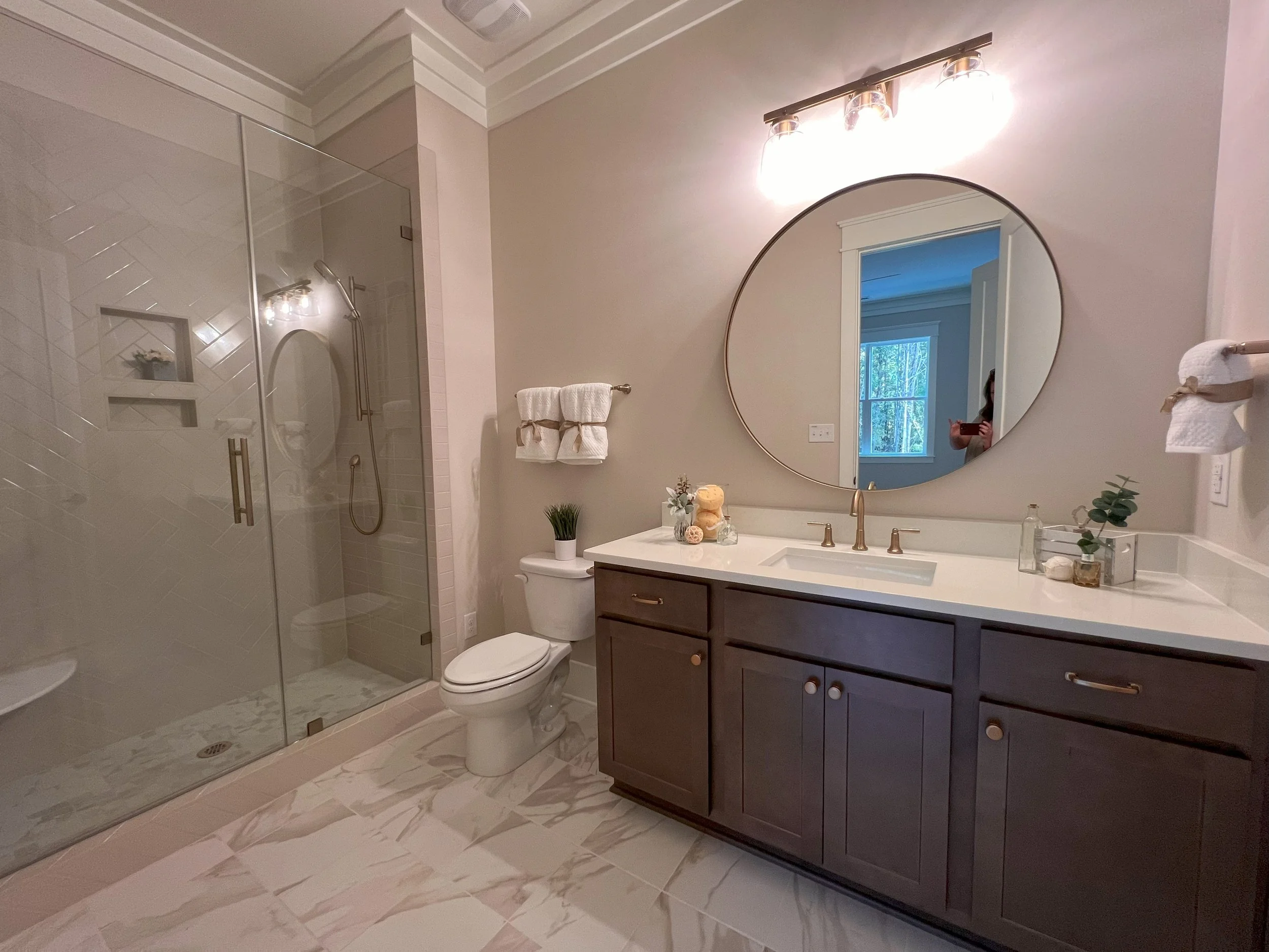 Modern bathroom with a glass-enclosed shower on the left, a toilet in the middle, and a dark wood vanity with a white countertop on the right. Above the vanity is a large round mirror, and a light fixture with three bulbs. Decor includes small plants