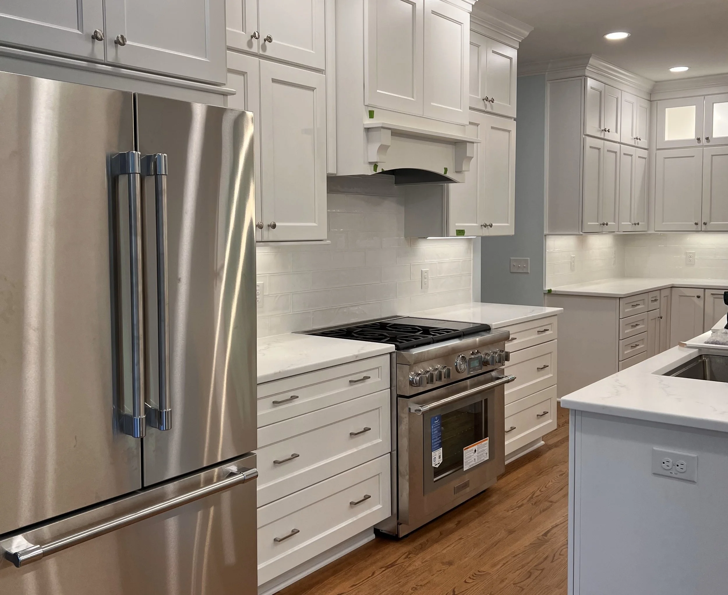 Modern kitchen with white cabinets, marble countertops, a stainless steel refrigerator, and a stove with a hood, wood floors, and multiple electrical outlets on the walls.