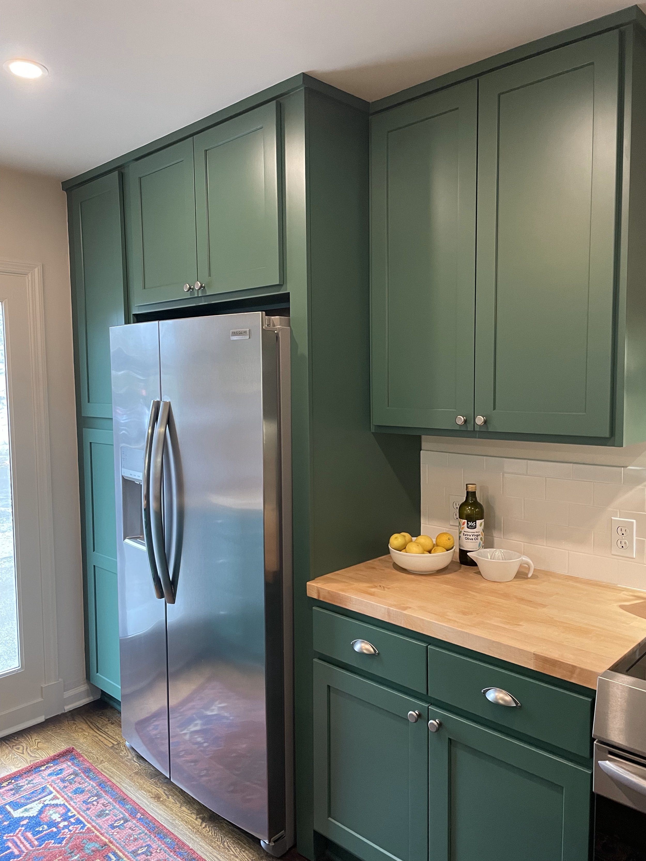 Kitchen corner with green cabinets, a stainless steel refrigerator, a wooden countertop with a bowl of lemons, a bottle of olive oil, and a white mixing bowl.