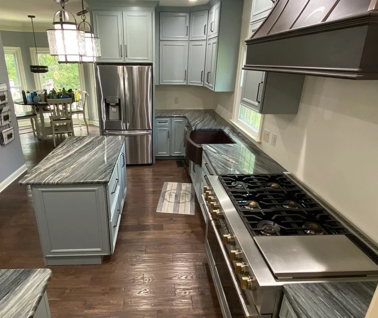 A modern kitchen with gray cabinets, stainless steel appliances, a marble countertop island, a dark brown sink, a gas stove, and hardwood flooring. There is a dining area visible in the background with a table and chairs.