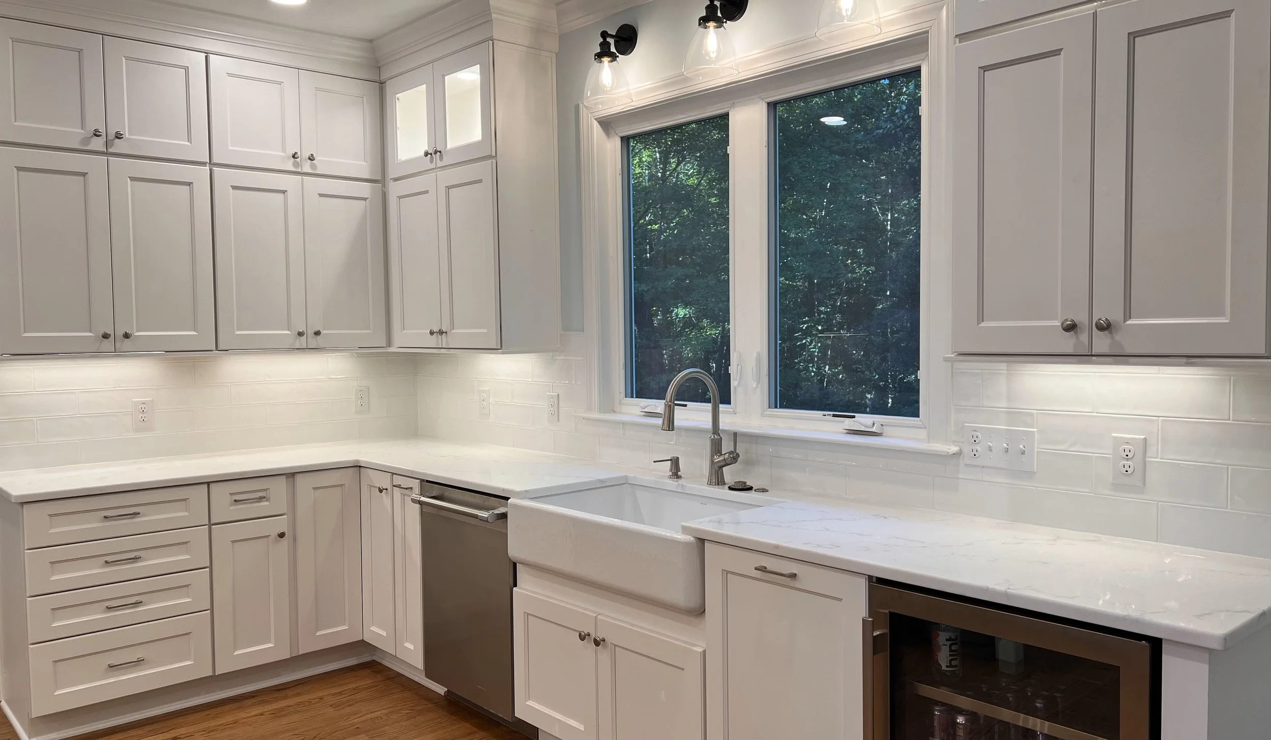 Modern white kitchen with upper and lower cabinets, white marble countertops, a farmhouse sink under a large window, and stainless steel appliances. Recessed and sconce lighting, hardwood floor, and lush greenery outside the window.