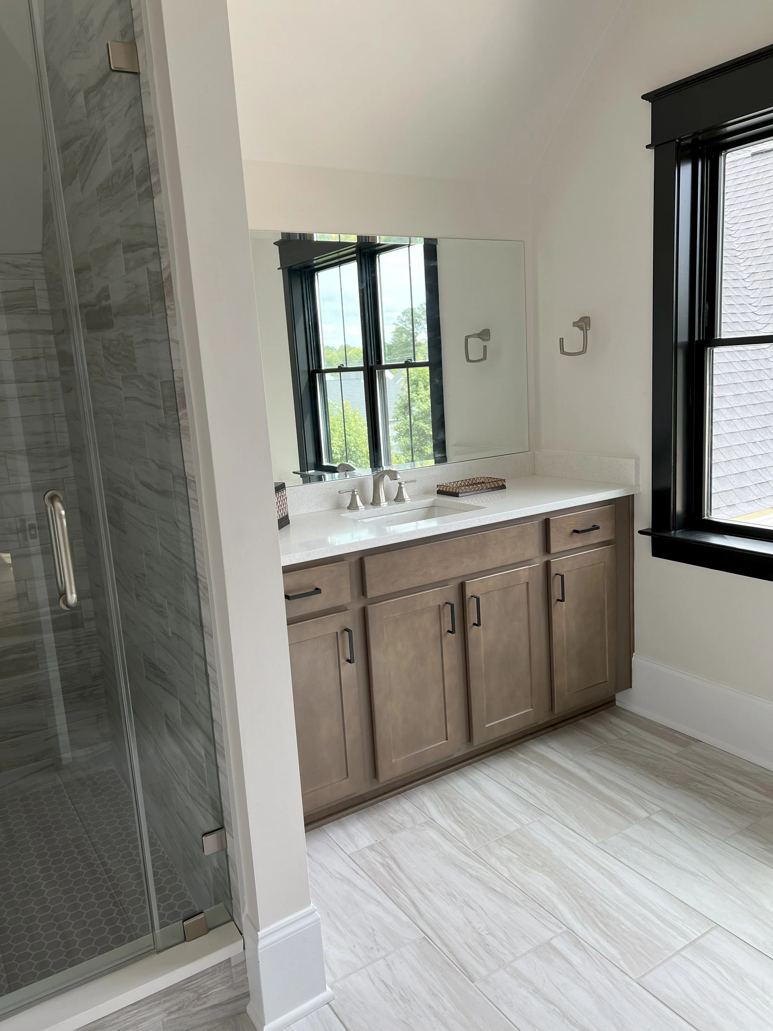 Bathroom vanity area with a large mirror, window, wooden Cabinet, stack of towels, and a shower with glass door and tiled wall.