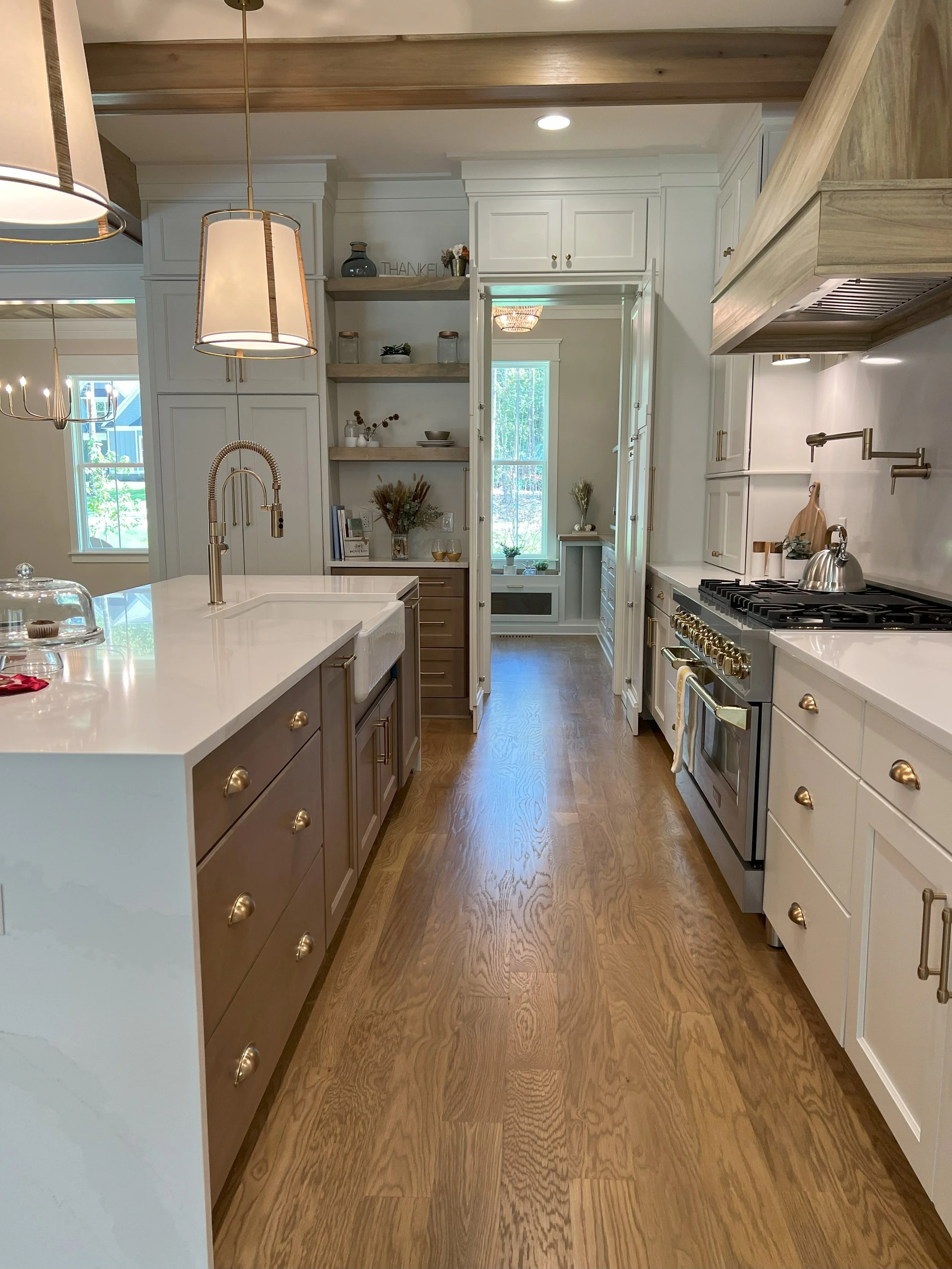 Modern kitchen with white and beige cabinets, wooden floor, central island with sink, gold hardware, and a stove with gold knobs. Pendant lights hanging over the island, open shelves, and windows providing natural light.