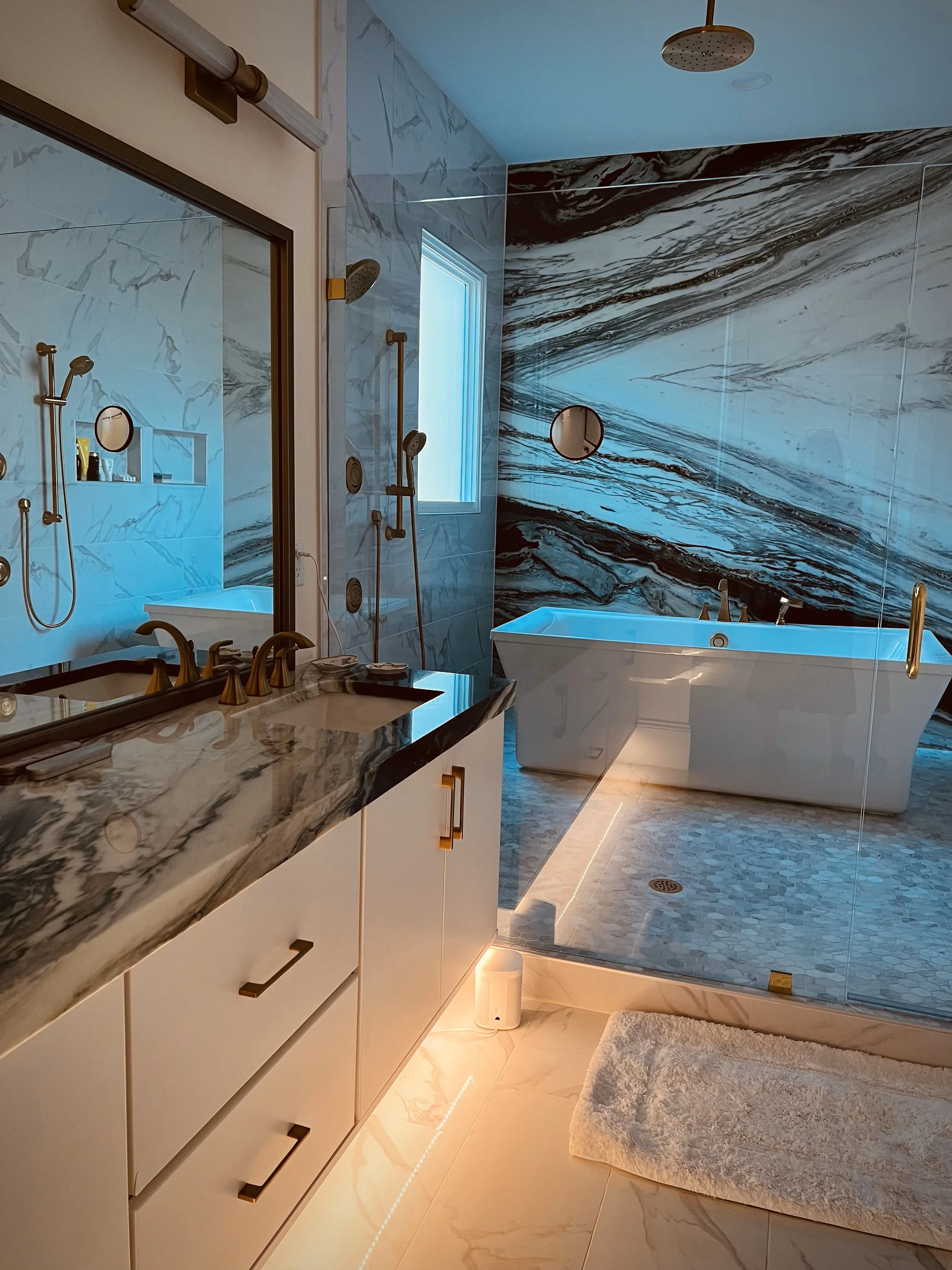 Modern bathroom featuring a marble countertop vanity with gold hardware, a large mirror, a relaxing bathtub in front of a marble accent wall, a walk-in shower with multiple showerheads, a window providing natural light, and soft lighting under the va
