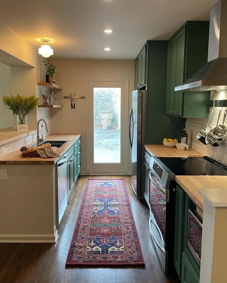 A cozy kitchen with green cabinets, wooden countertops, and a patterned red runner rug. There's a door at the end leading outside, with a small shelf, flowers, and utensils on the left, and kitchen appliances including a stove, microwave, and refrige