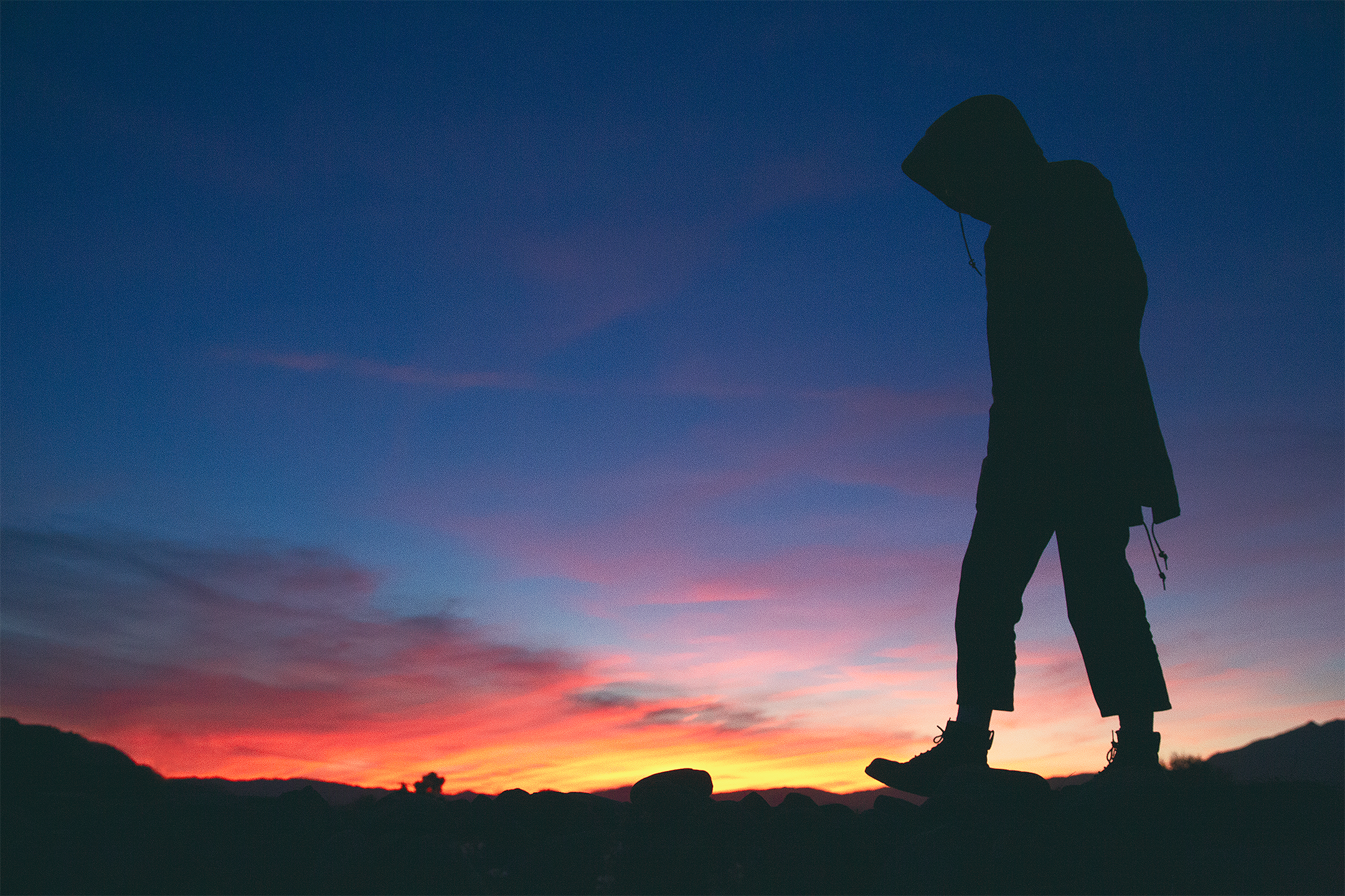 Silhouette of a person walking on rocky terrain during sunset with a colorful sky and distant mountains.