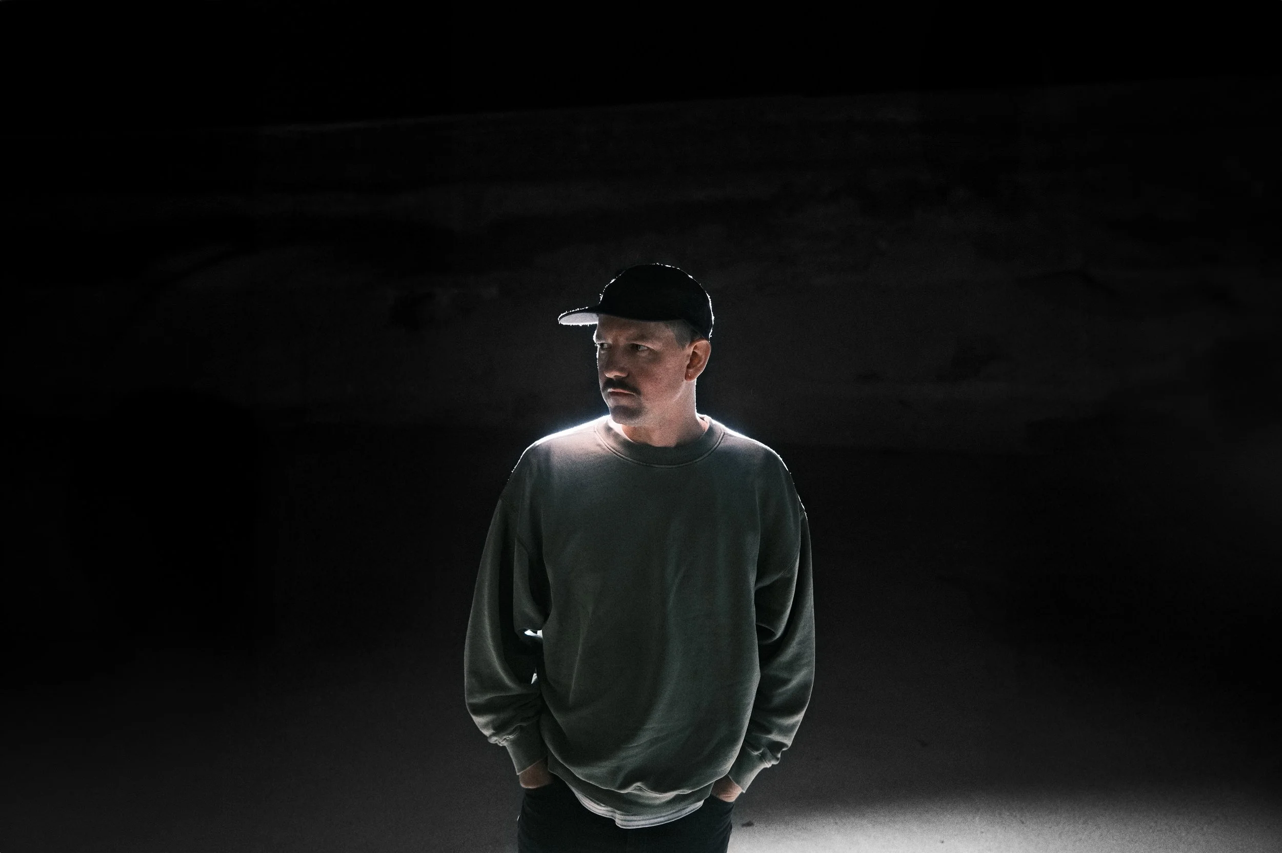 A man with a mustache wearing a cap and a gray sweatshirt, standing in a dark environment with a light source behind him.