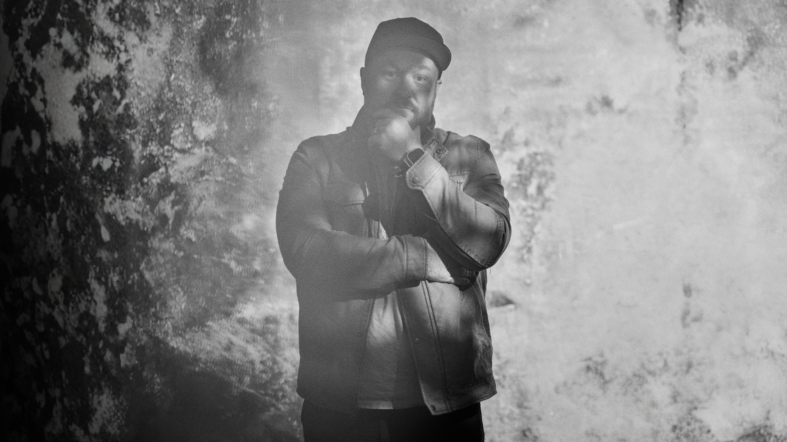 A man in a leather jacket and beanie hat standing with one arm crossed and his other hand touching his chin, against a background that appears to be a textured wall or natural scenery, in black and white.