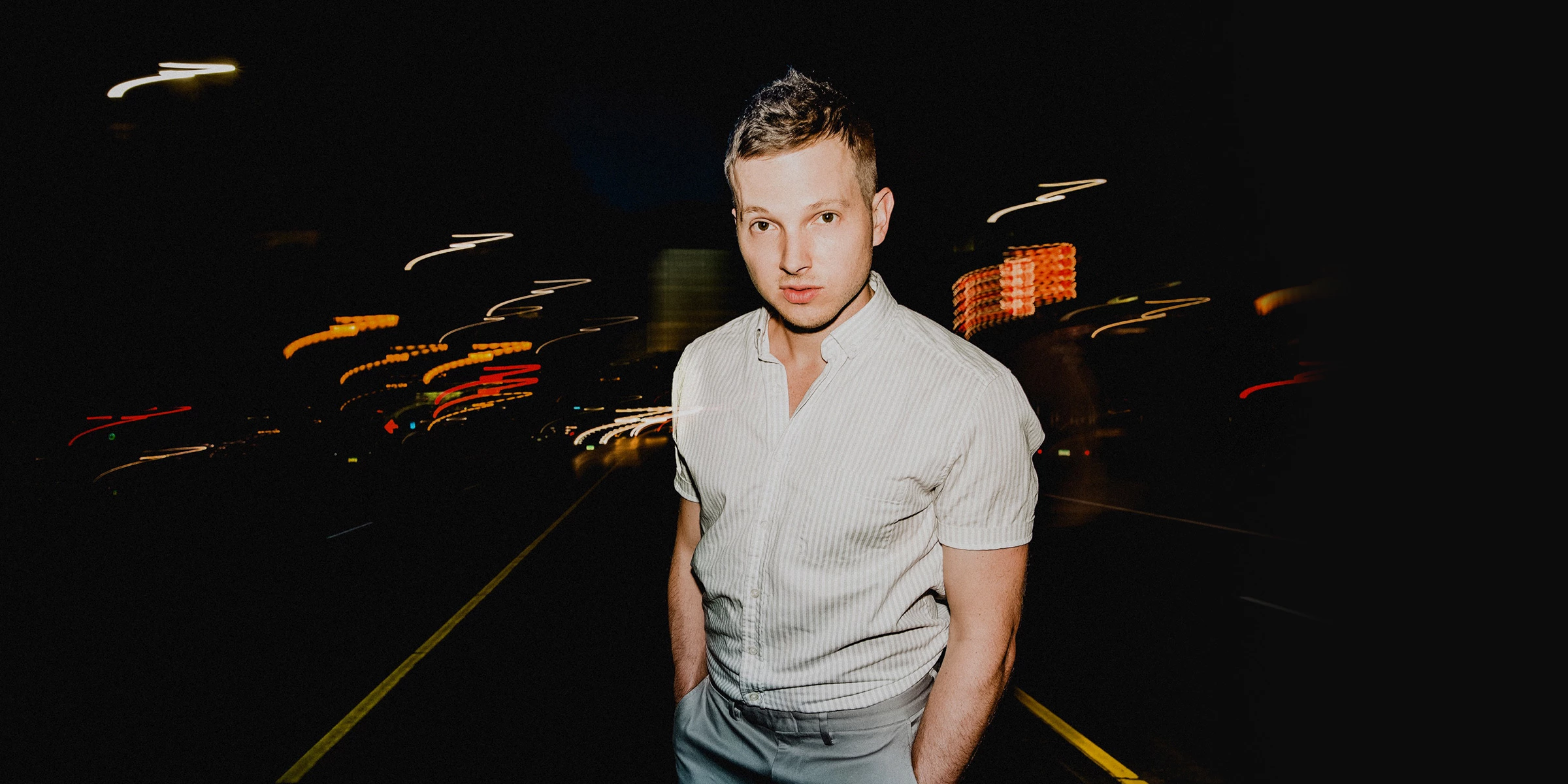 A young man with short hair wearing a light-colored, short-sleeved, button-up shirt and gray pants standing in the middle of a dark street at night with blurred city lights behind him.