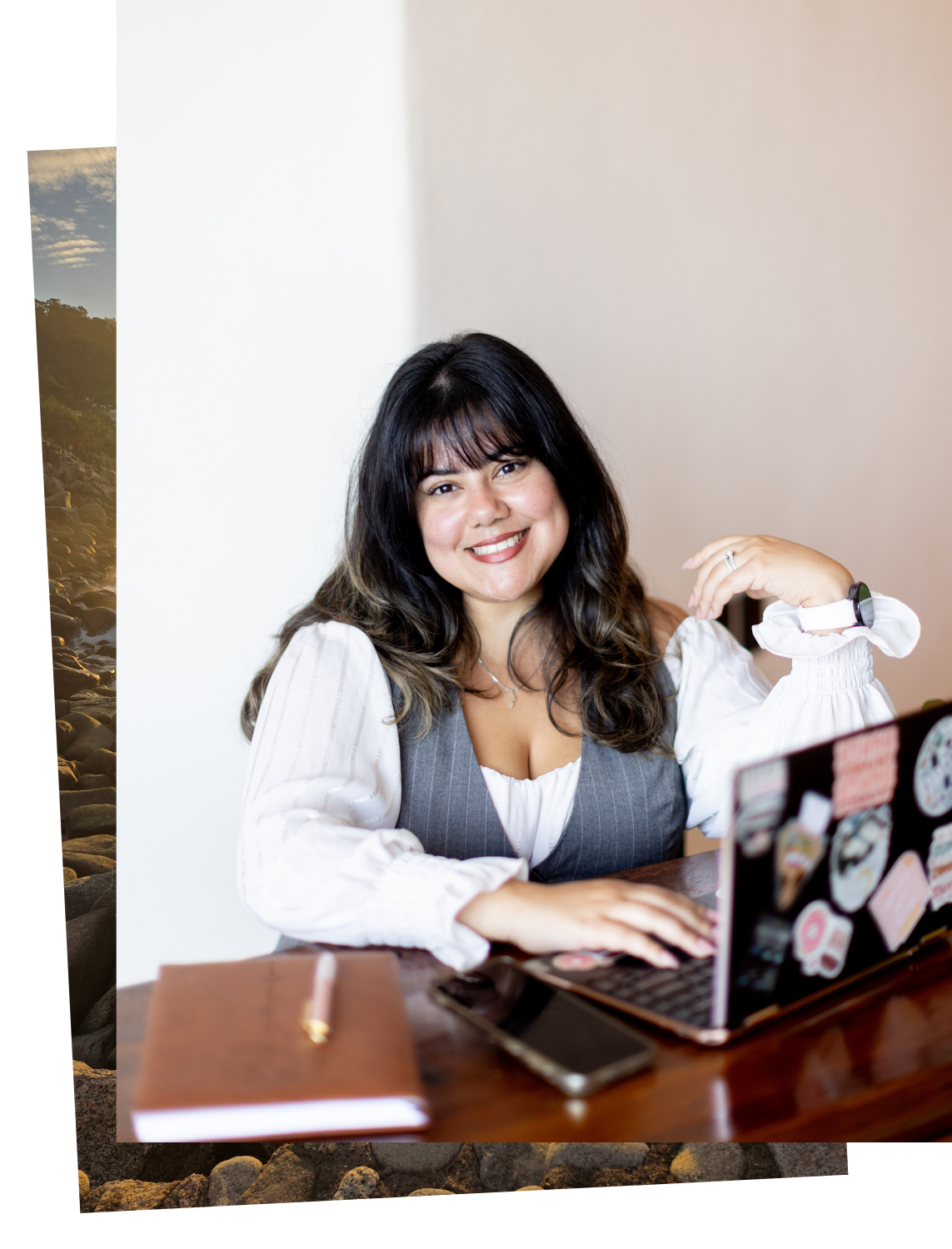 A smiling woman with dark hair and bangs sitting at a wooden desk with a laptop covered in stickers, a smartphone, a brown notebook, and a pen.