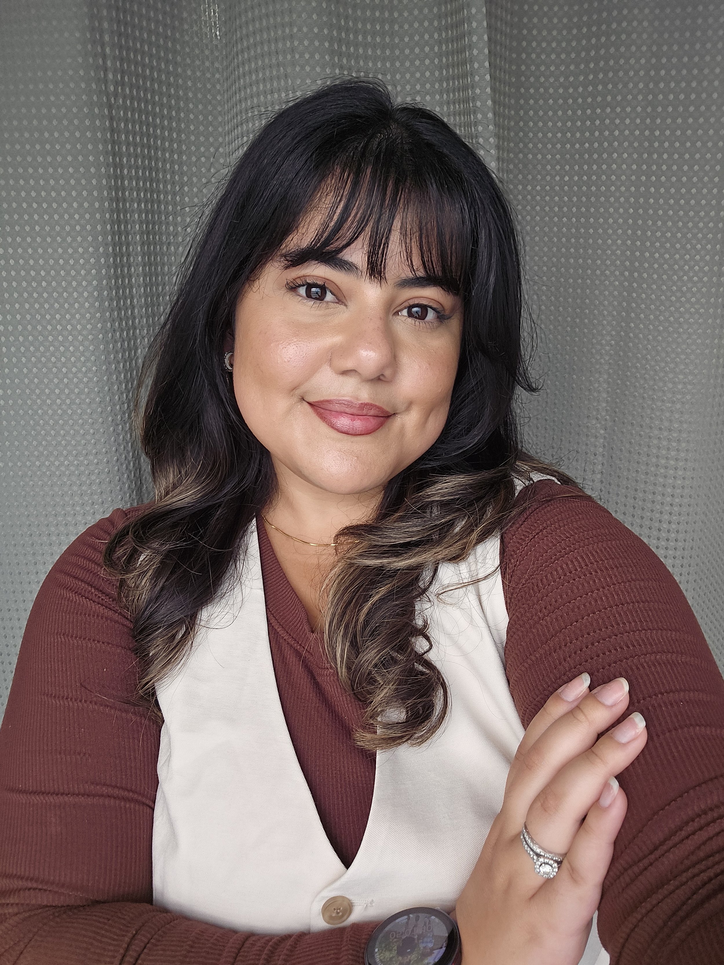 A woman with shoulder-length dark brown hair with lighter tips, wearing a maroon long-sleeve shirt and a cream-colored vest, smiling and looking at the camera in front of a gray perforated metal background.