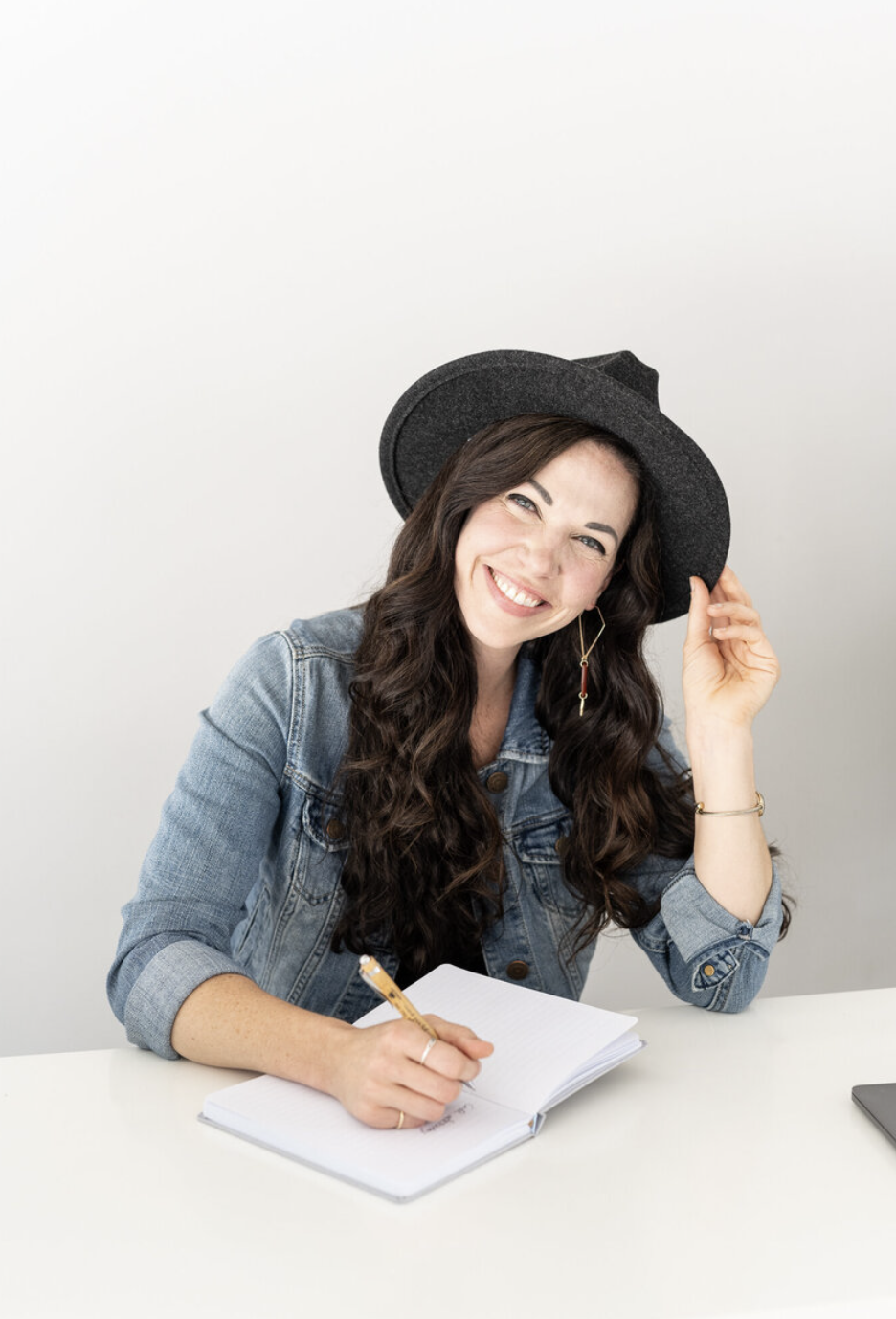 A woman with long dark hair, smiling, sitting at a white desk with a notebook, wearing a denim jacket, hoop earrings, a gold bracelet, and a black wide-brimmed hat.