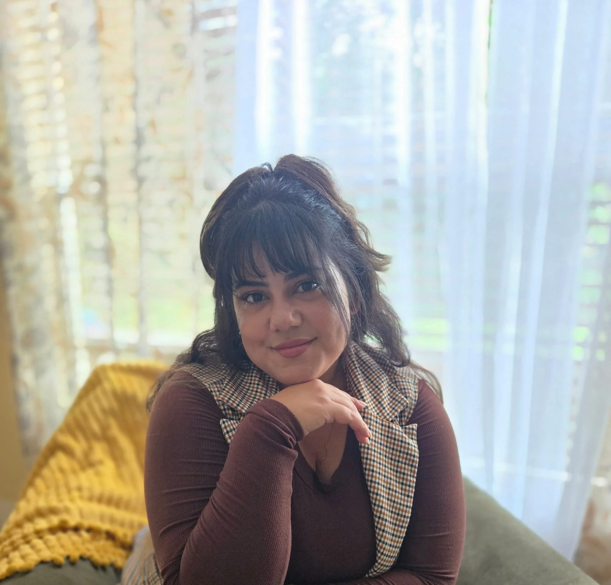 A woman with dark, wavy hair and bangs, sitting on a green couch with yellow pillows, resting her chin on her hand, in front of sheer curtains with trees visible outside.