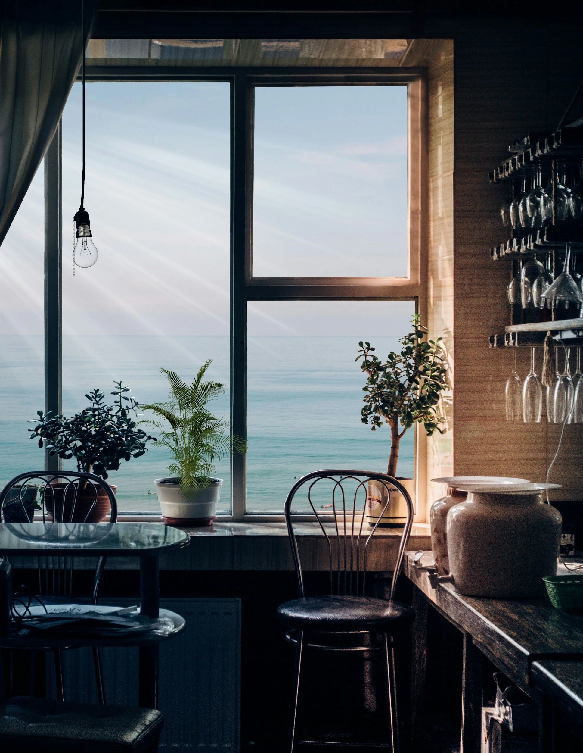 Inside a cozy restaurant with large ocean view window, potted plants on windowsill, hanging glasses, and wooden decor.